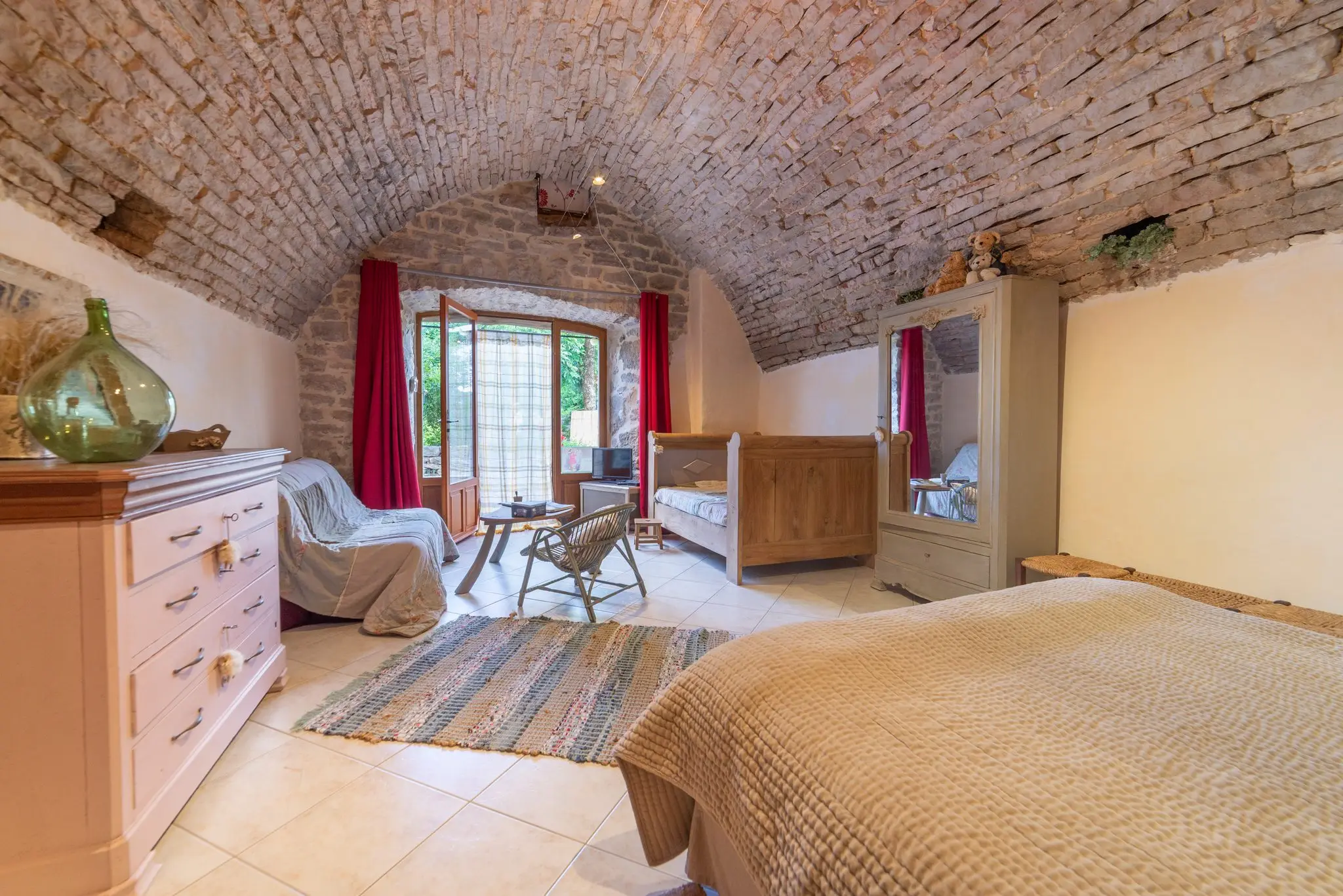 Chambre sous voûte en pierre avec lit double au premier plan, tapis tissé, lit de bois et porte-fenêtre encadrée de rideaux rouges