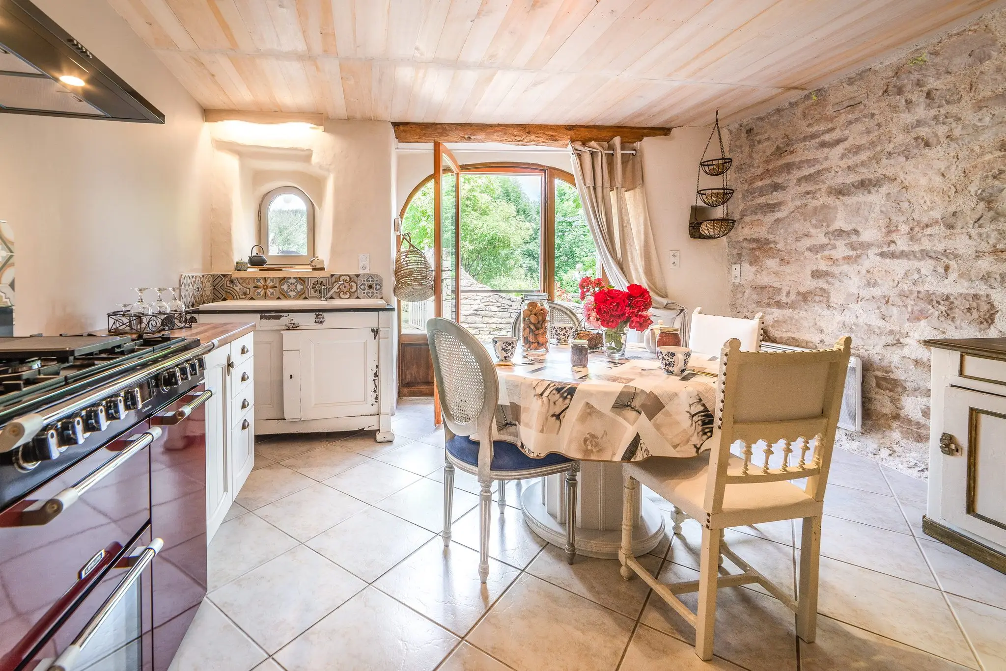 Cuisine au piano de cuisson bordeaux, table ronde dressée avec un bouquet de roses, mur en pierre et porte-fenêtre cintrée ouverte sur la verdure