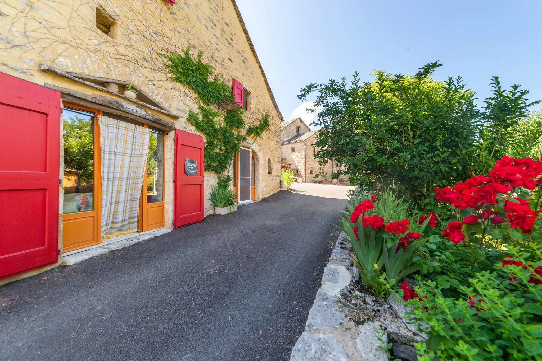 Porte vitrée aux volets rouges, rideau à carreaux et massif de rosiers rouges au pied d'une façade en pierre de hameau