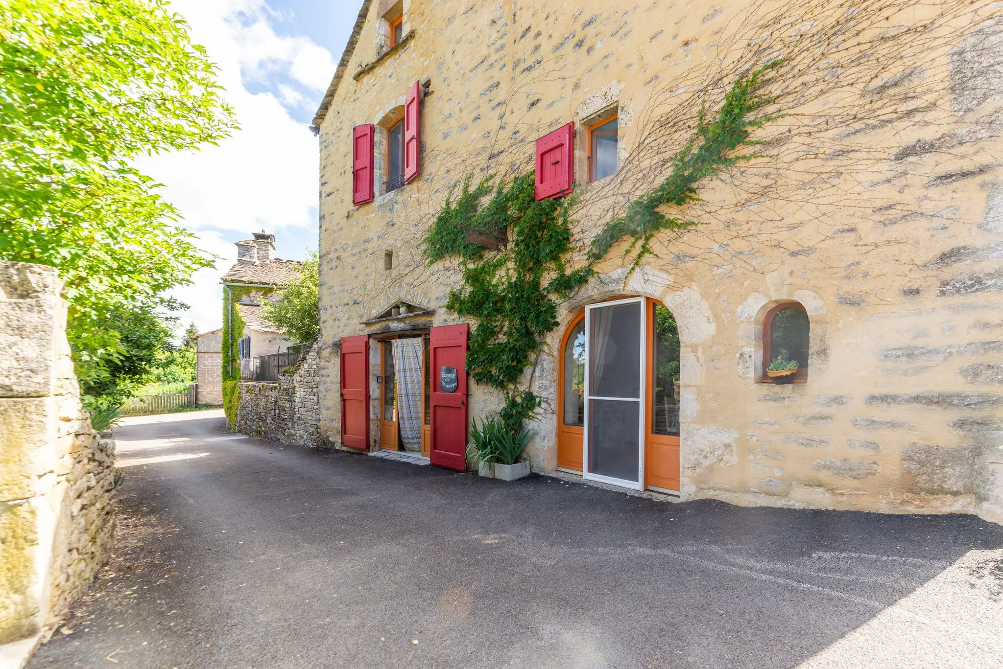 Longue façade en pierre blonde d'une ancienne bergerie, volets rouges, portes vitrées orange et vigne grimpante le long d'une ruelle de hameau