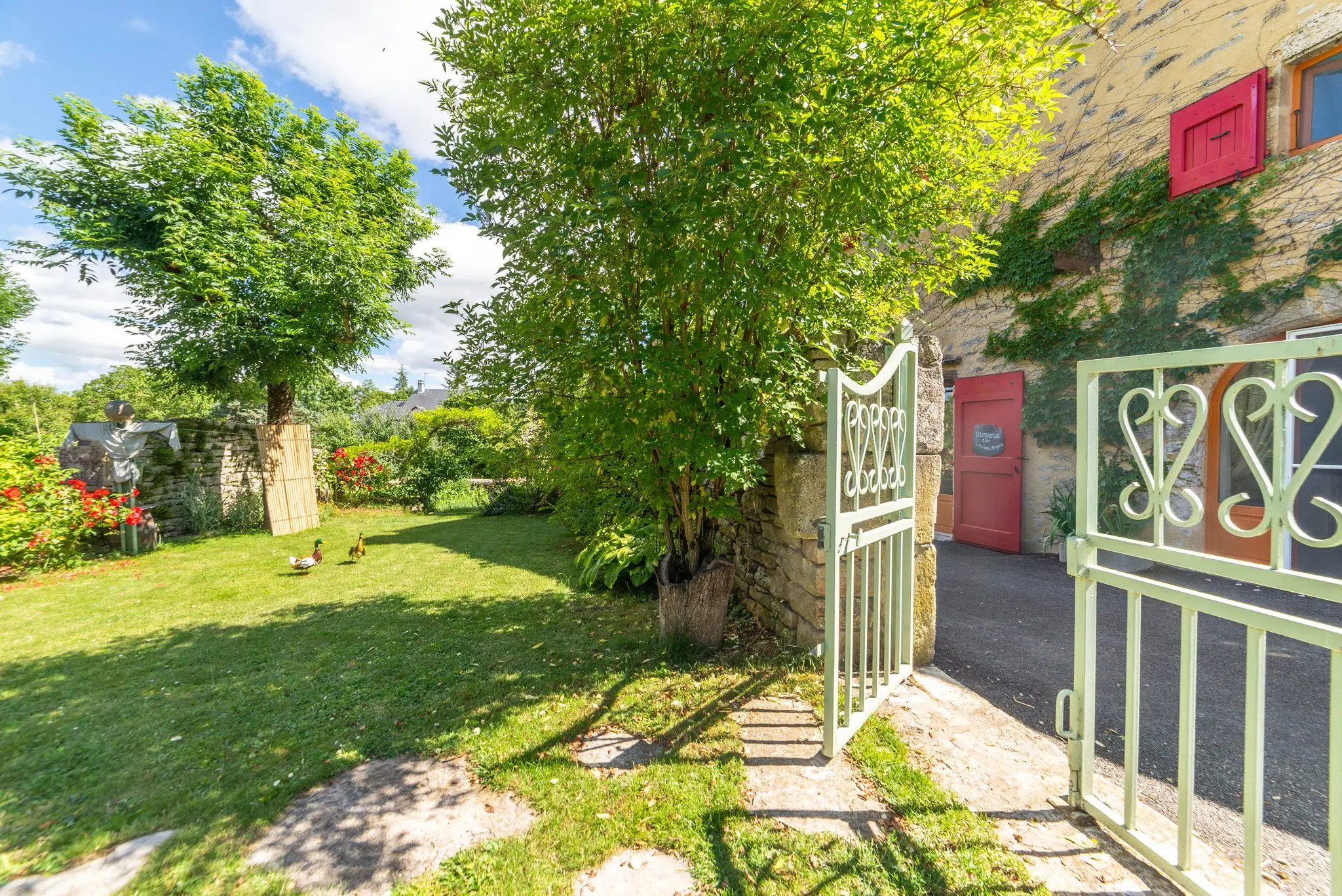 Portillon en fer forgé vert d'eau ouvert sur une pelouse ombragée d'arbres, à côté d'une maison en pierre aux volets rouges