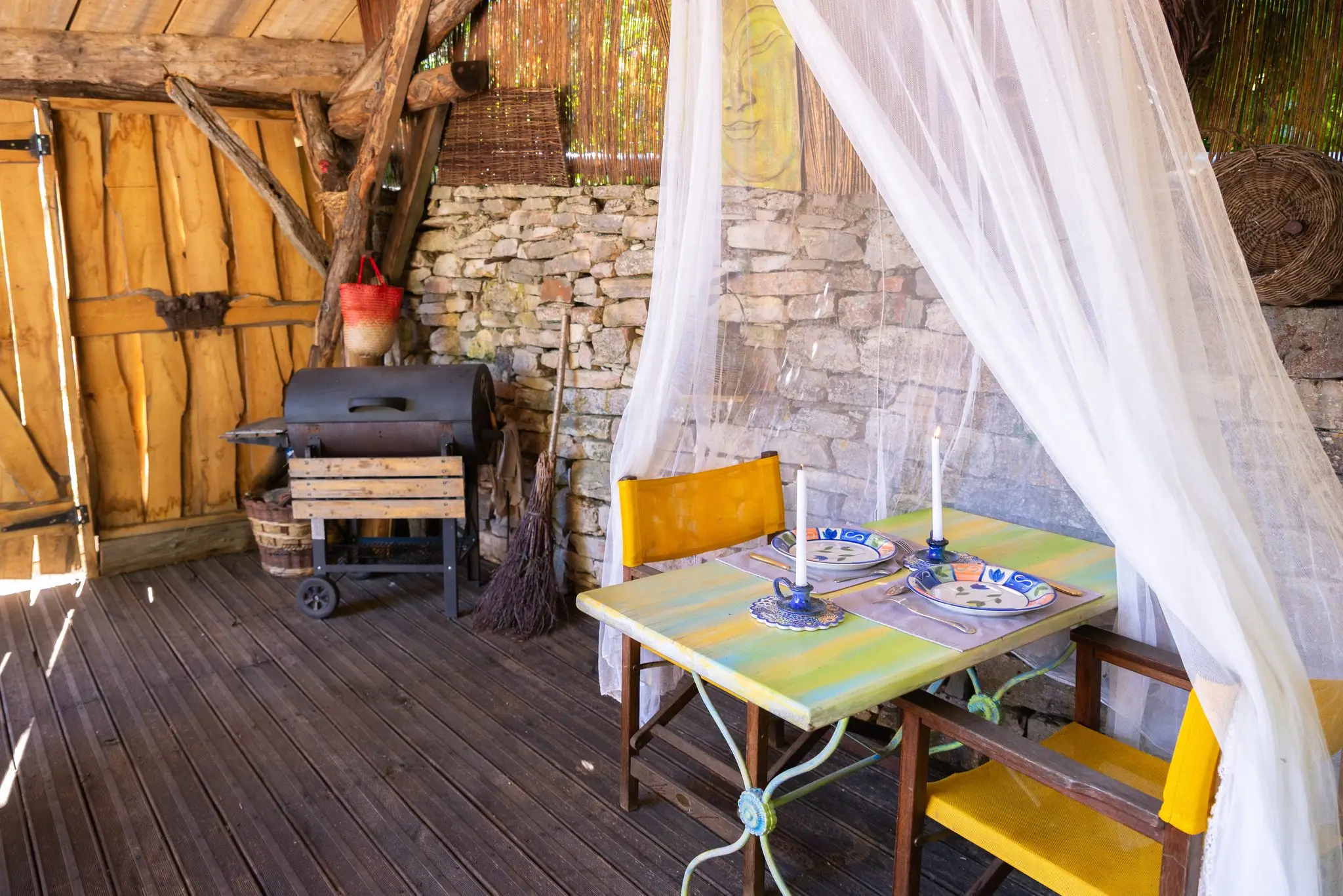 Petite table dressée de bougies sous un voilage blanc, chaises jaunes, barbecue et muret en pierre sous l'abri en bois