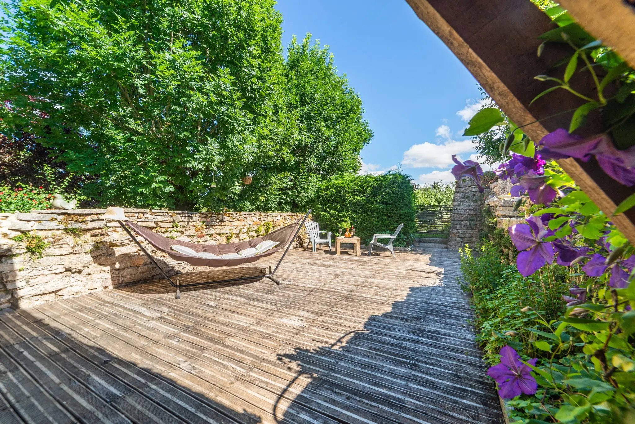 Terrasse en lames de bois bordée d'un muret en pierre sèche, hamac, fauteuils de jardin blancs et clématite violette au premier plan