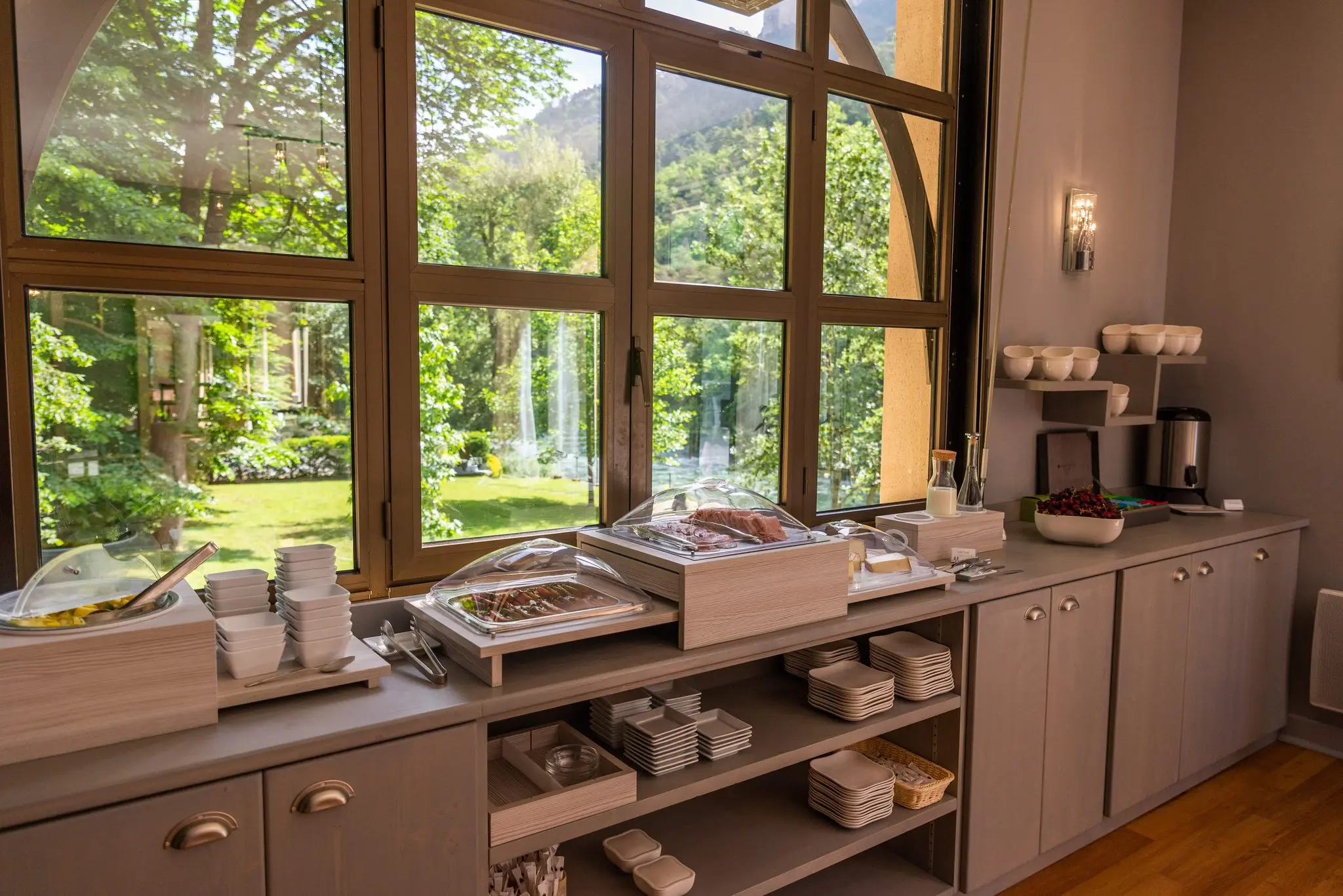 Buffet de petit-déjeuner dressé le long d'une grande baie vitrée cintrée donnant sur le jardin ensoleillé
