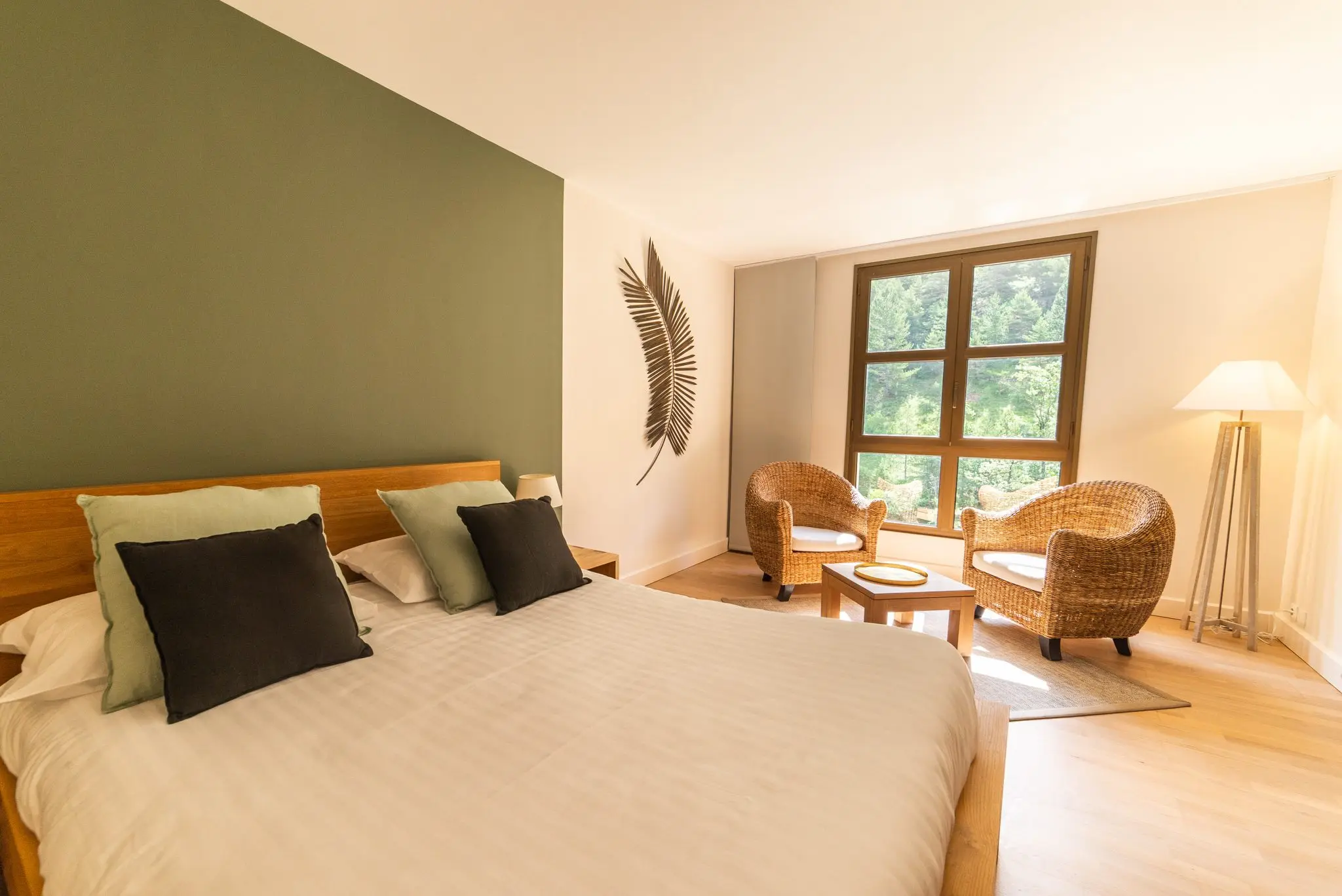 Chambre d'hôtel au mur vert sauge, coussins assortis et deux fauteuils en rotin devant la fenêtre