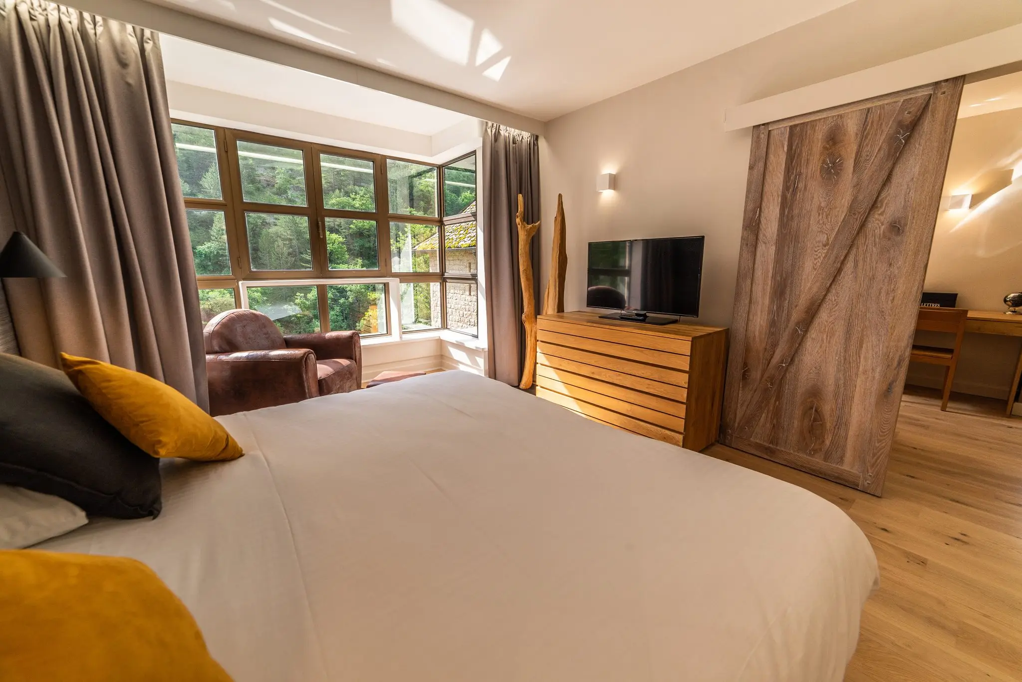 Chambre d'hôtel lumineuse, porte coulissante en bois brut, fauteuil club et vue sur la forêt