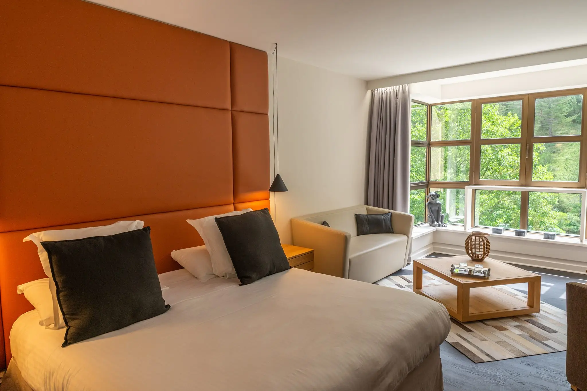Chambre avec haute tête de lit capitonnée orange, coin salon et bow-window ouvert sur la forêt