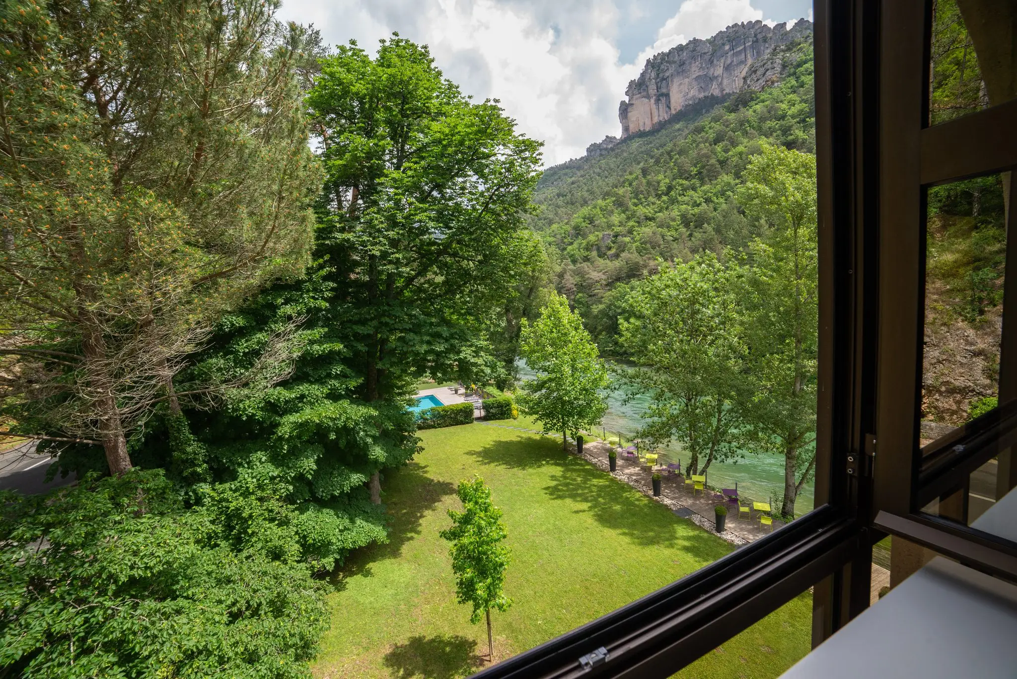 Pelouse arborée descendant vers le Tarn, piscine entre les arbres et falaise calcaire au fond, vue depuis une fenêtre d'étage