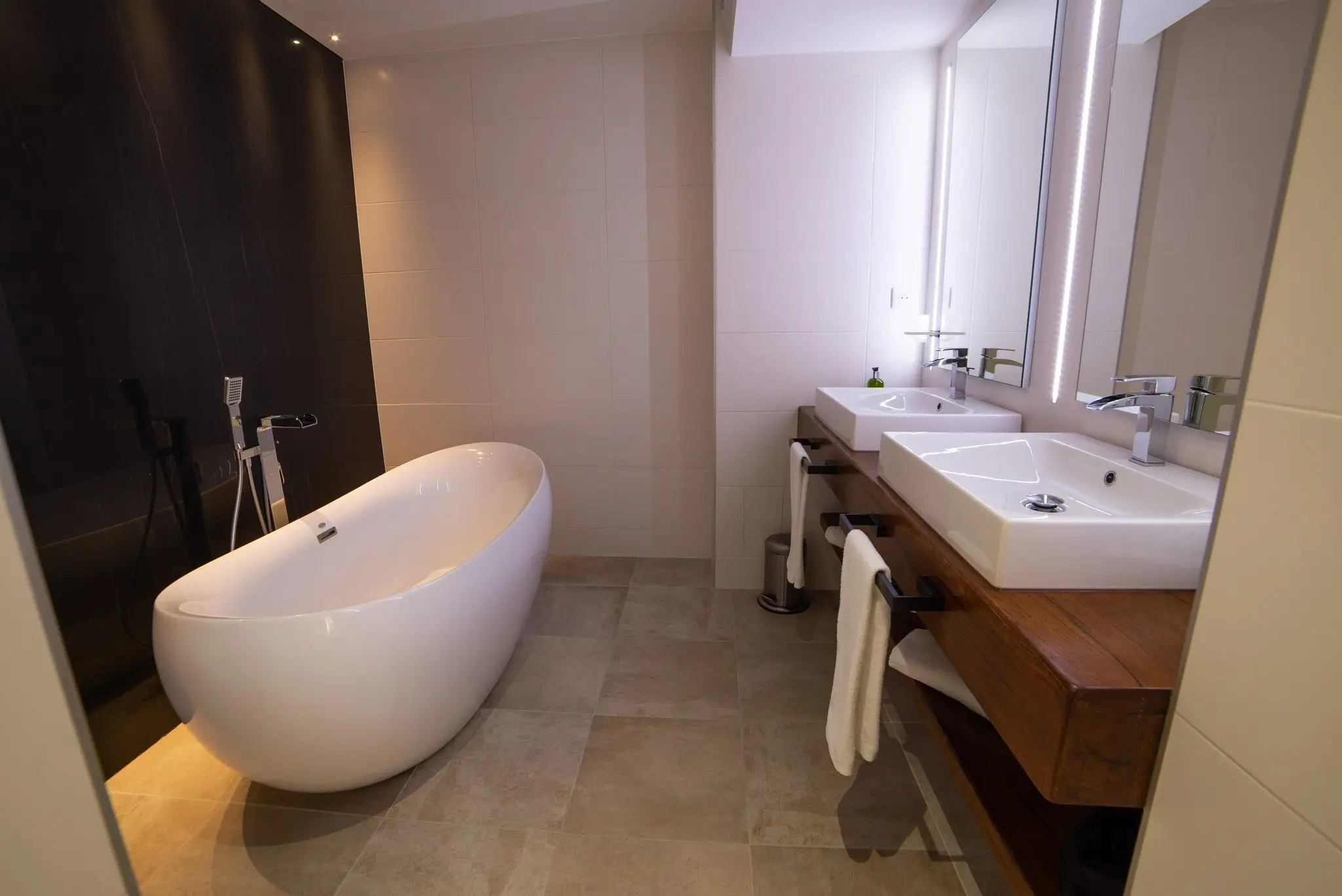Salle de bains à baignoire îlot blanche devant un mur noir, double vasque sur plan en bois et grand miroir
