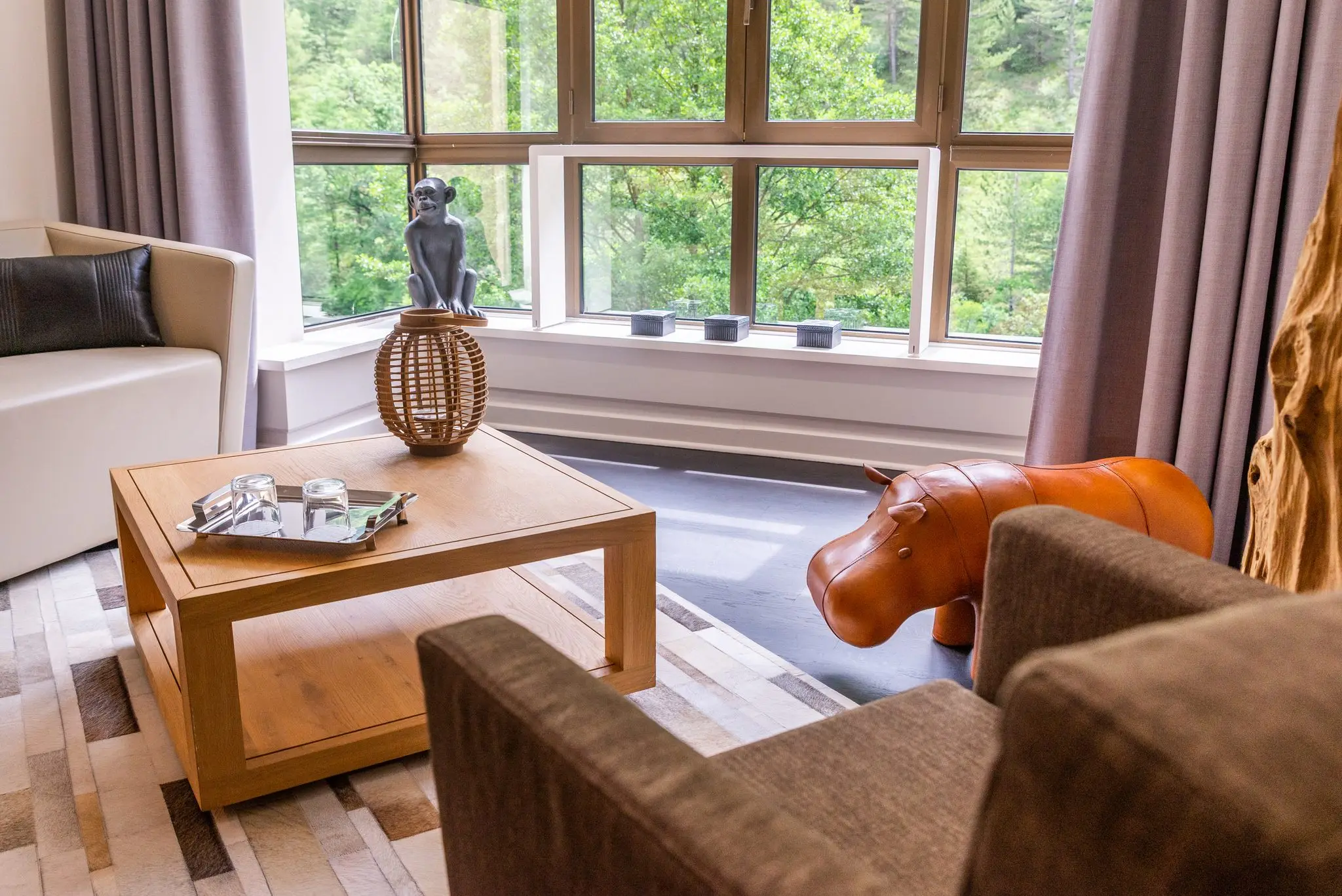 Salon d'hôtel avec baie vitrée sur la forêt, table basse en bois et pouf en cuir en forme d'hippopotame