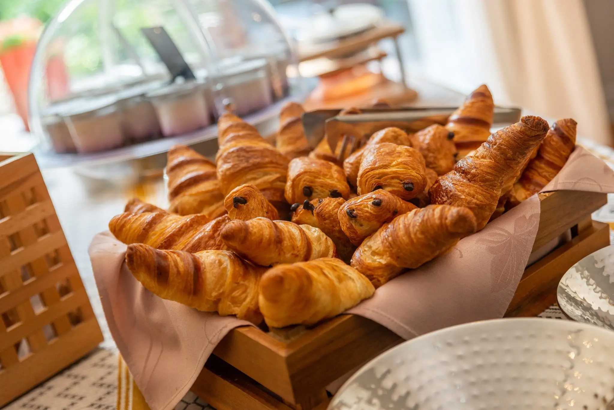 Corbeille de croissants et de pains au chocolat dorés posée sur le buffet, cloche à yaourts en arrière-plan flou