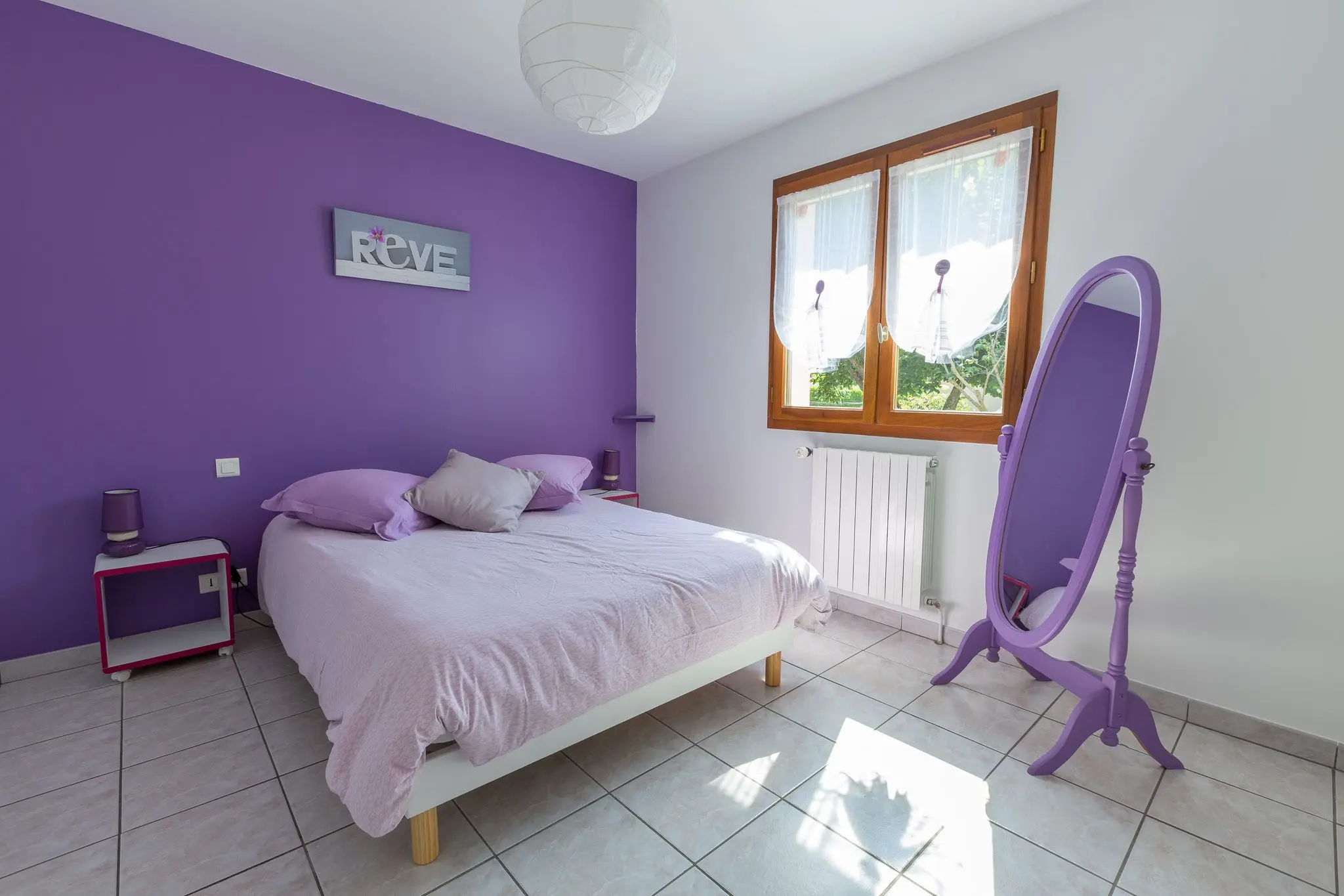 Chambre au mur d'accent violet, lit double, psyché mauve et fenêtre à voilages, sol carrelé