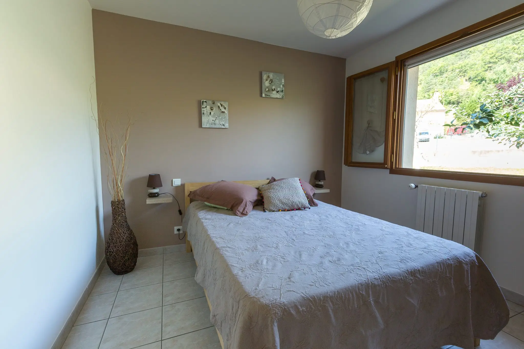 Chambre sobre au mur taupe, lit double, vase de branchages et large fenêtre sur la colline boisée