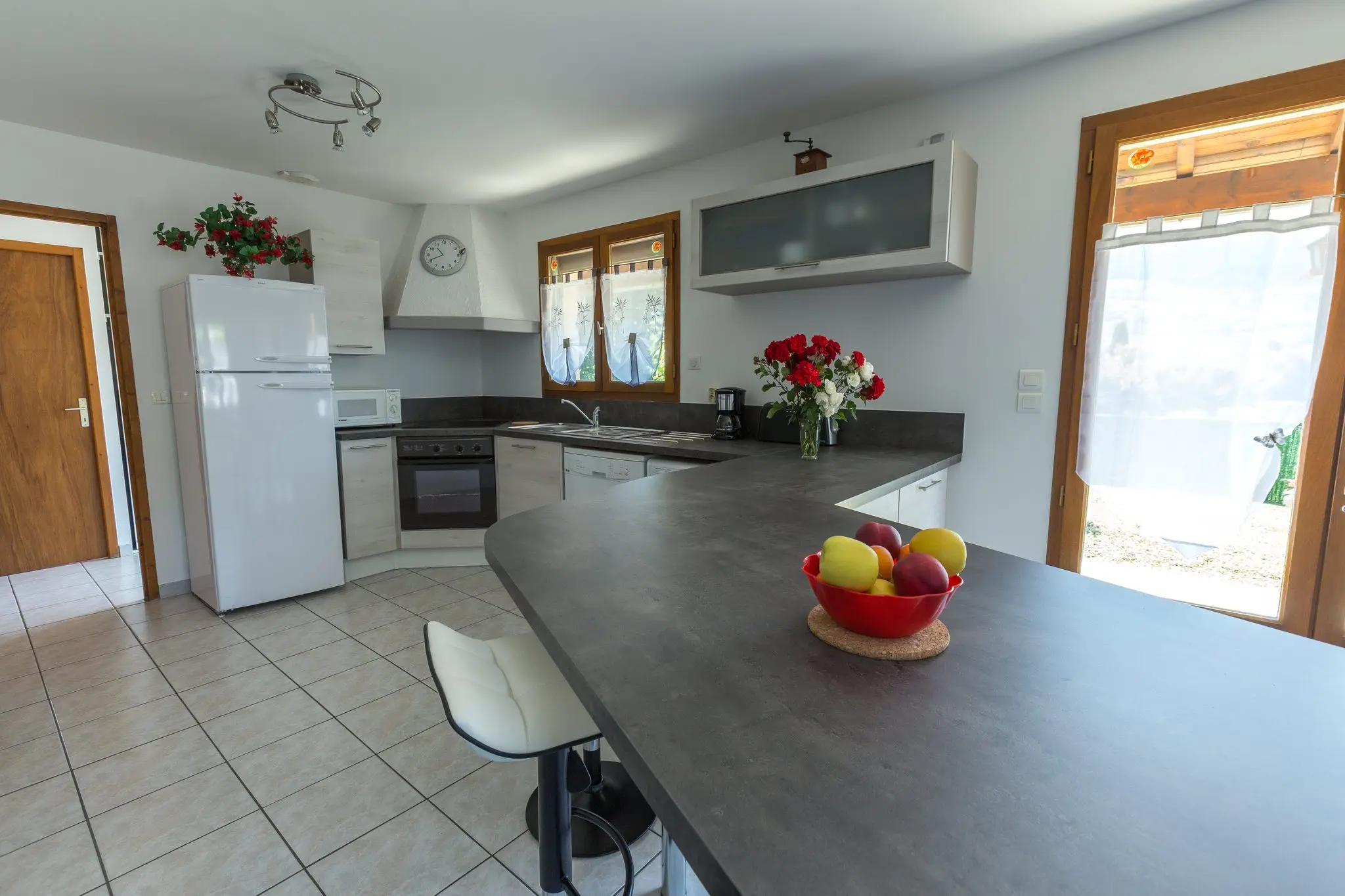 Cuisine équipée en L, plan de travail sombre prolongé d'un bar avec tabouret, réfrigérateur blanc et fenêtres sur le jardin