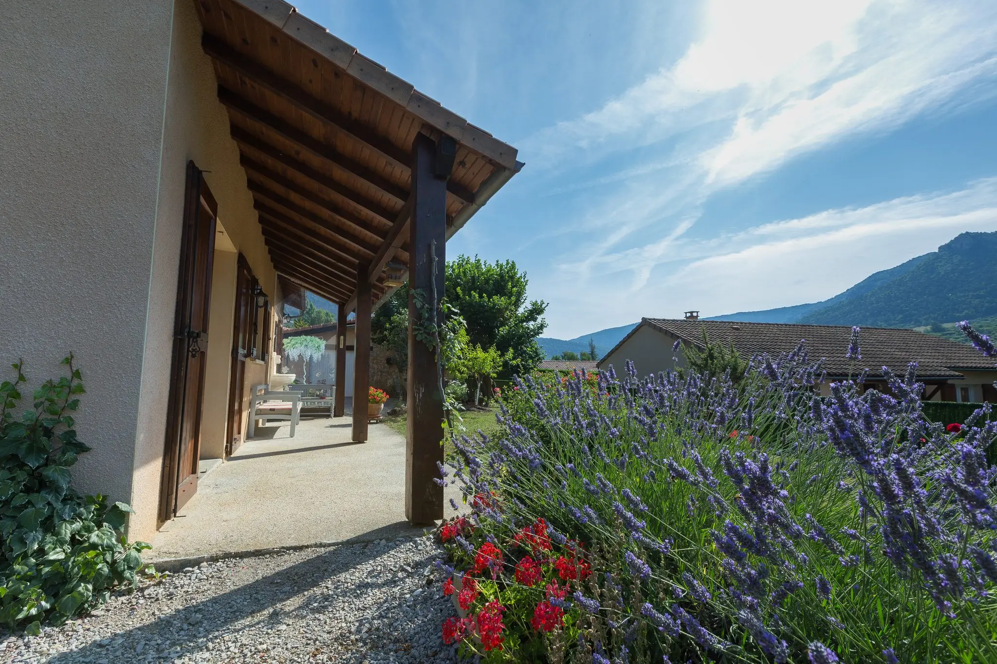Galerie couverte à charpente bois longeant la maison, bordure de lavande et géraniums rouges, montagnes au loin