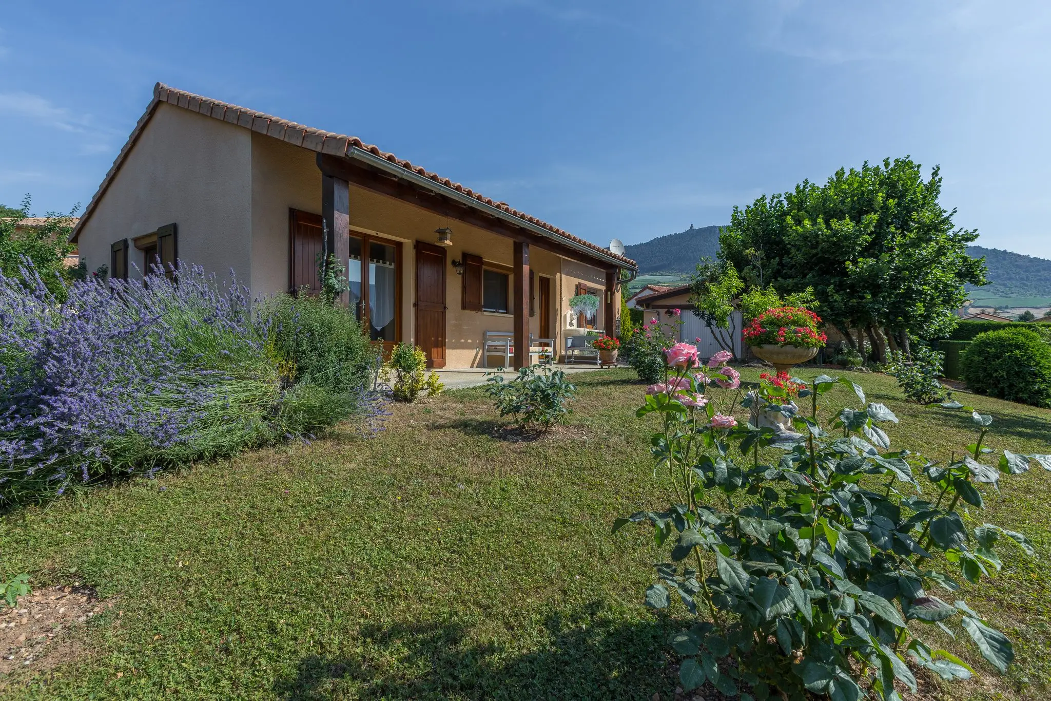 Maison de plain-pied aux murs crépis et toit de tuiles, galerie couverte, massifs de lavande et rosiers sur la pelouse, collines en arrière-plan