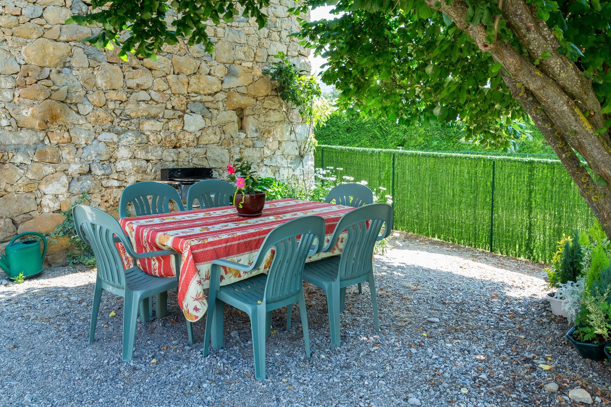 Table nappée et chaises de jardin sur gravier, sous un arbre, contre un ancien mur en pierres