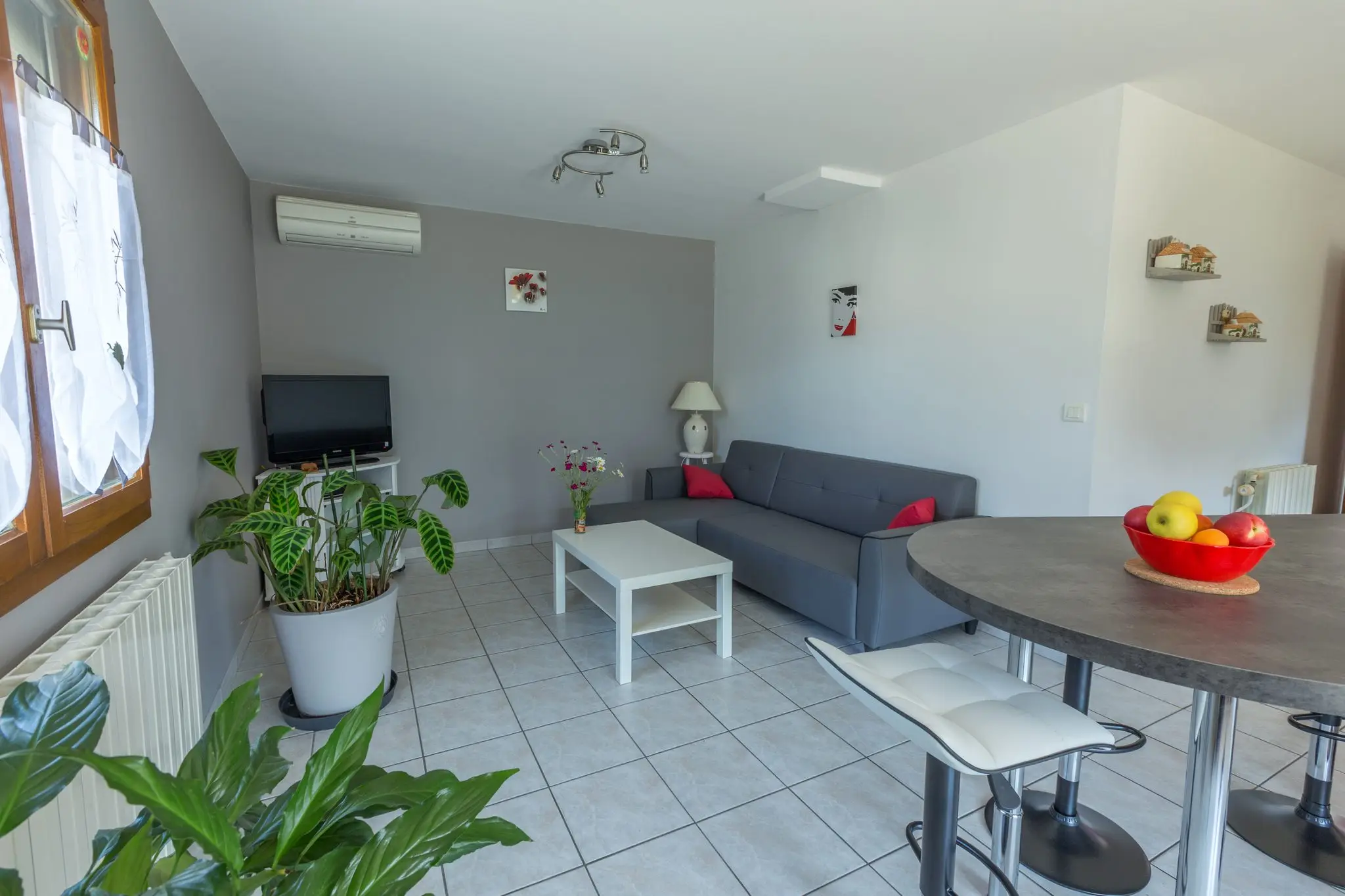 Séjour au carrelage clair, canapé gris, table haute et climatisation, grande fenêtre à voilages