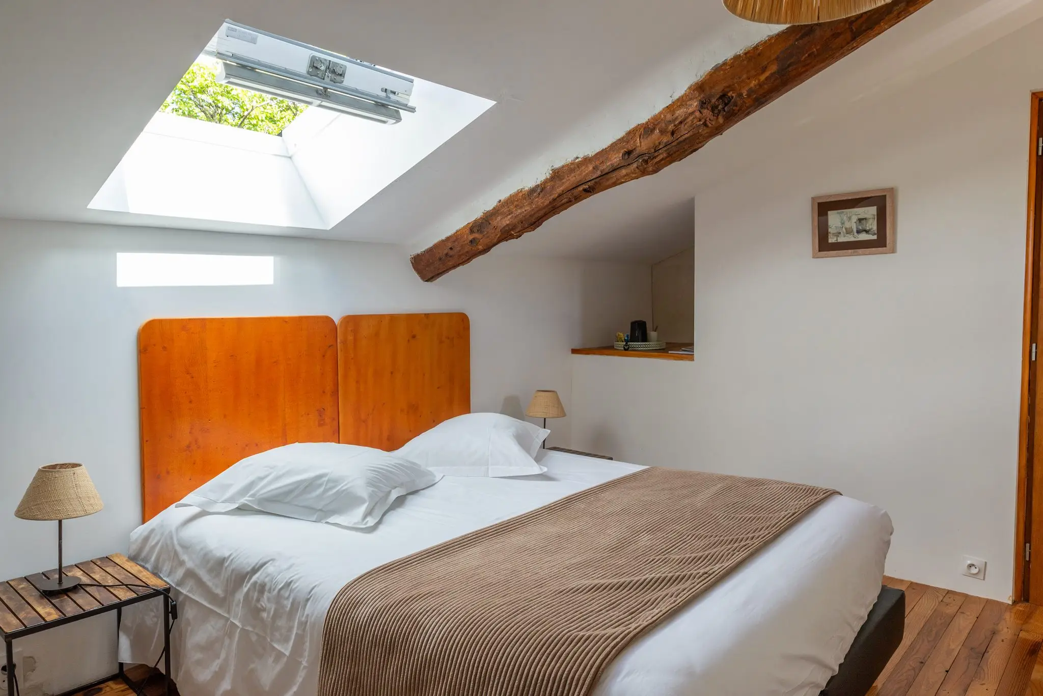 Chambre sous les combles, fenêtre de toit, poutre ancienne, tête de lit en bois et plaid brun sur un lit double
