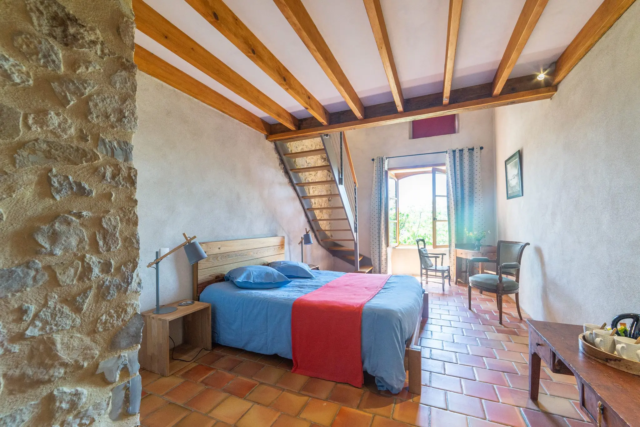Chambre sous poutres apparentes, mur en pierre brute au premier plan, lit à couvre-lit bleu et rouge, escalier de meunier vers la mezzanine et porte-fenêtre lumineuse