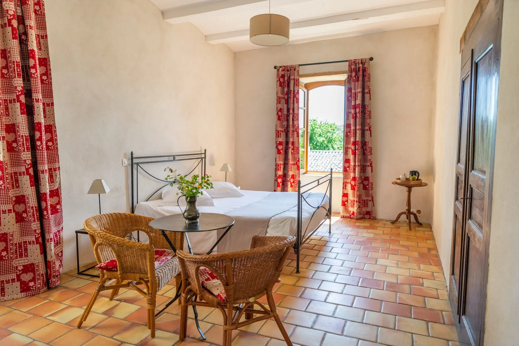 Chambre spacieuse au sol en tomettes, lit en fer forgé, fauteuils en rotin autour d'un guéridon fleuri, rideaux à motifs rouges et porte-fenêtre ouverte sur la verdure