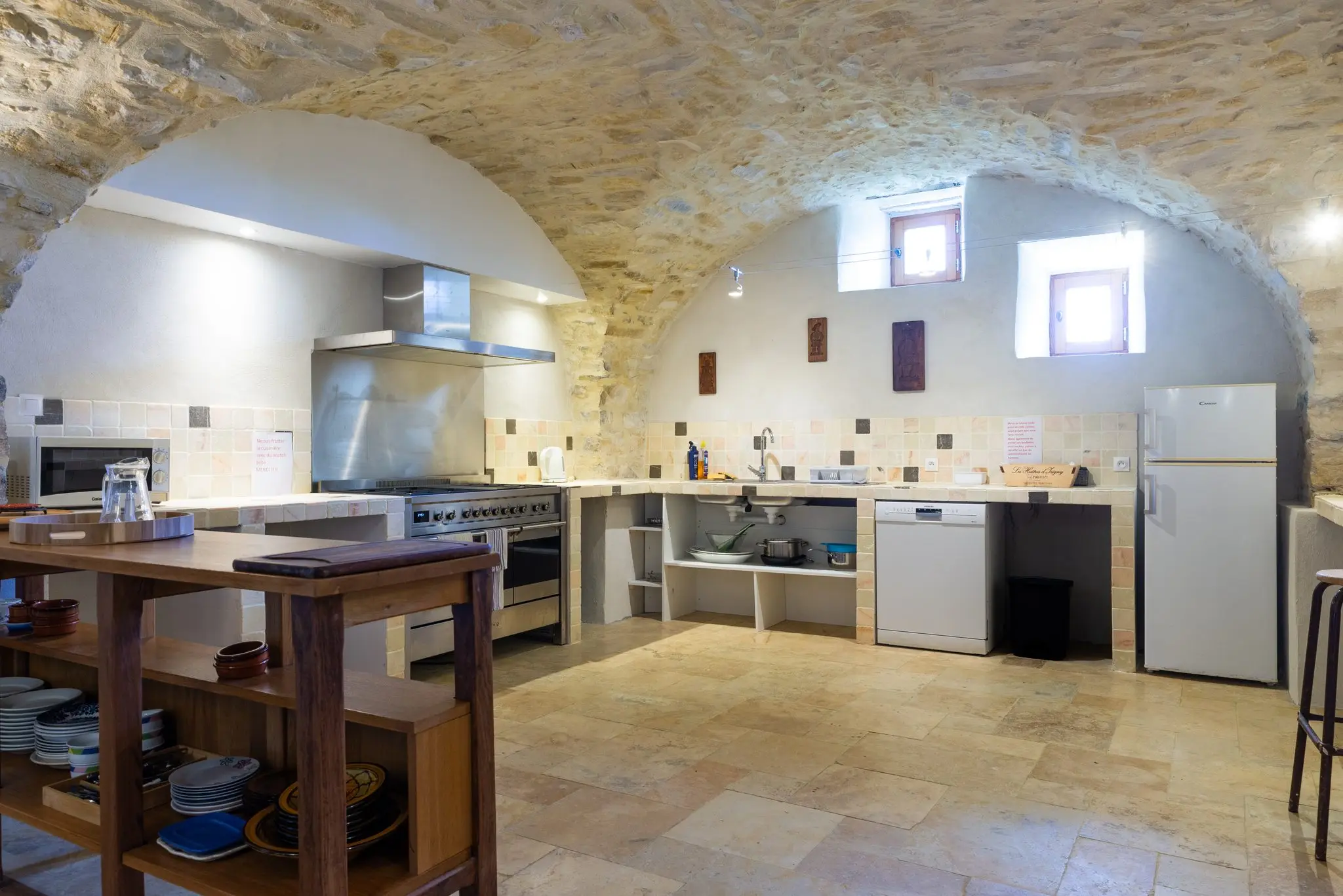 Cuisine équipée sous une voûte en pierre, piano de cuisson inox, îlot en bois, réfrigérateur et lave-vaisselle, sol en pierre claire