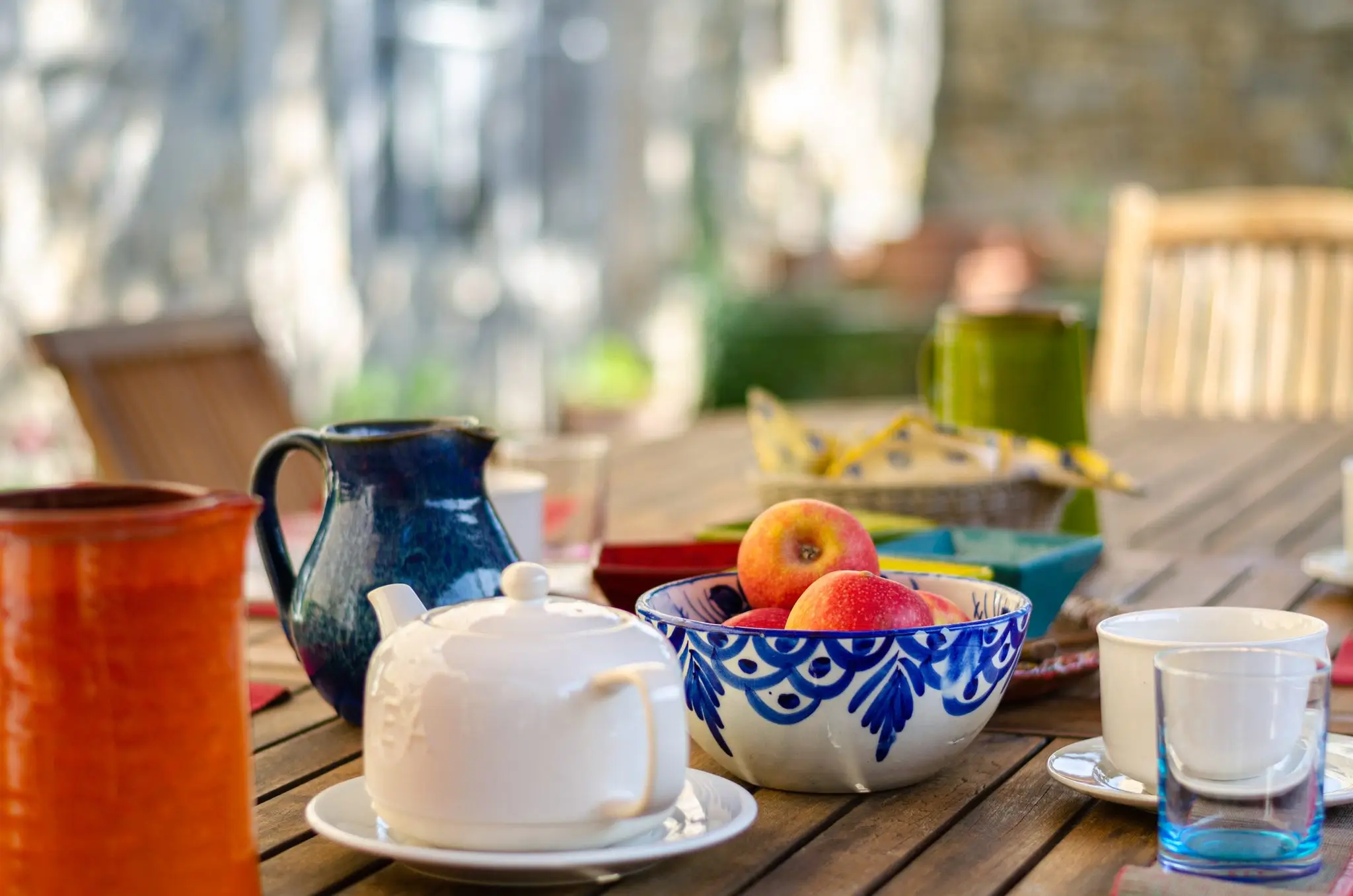 Théière blanche, pichets en céramique bleue et orange, bol de pommes et tasses sur une table en bois en extérieur