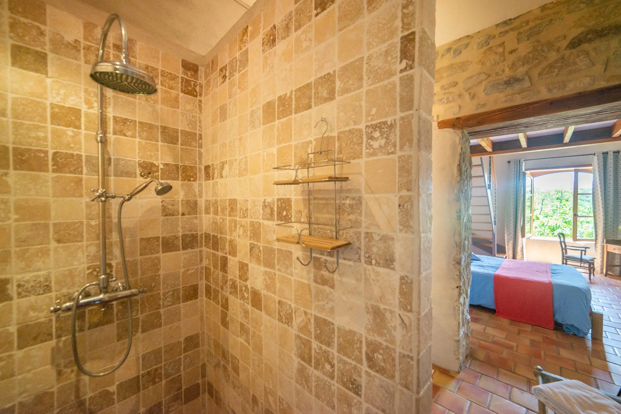 Douche à l'italienne carrelée de travertin avec colonne de douche chromée, ouverture vers une chambre à poutres et couvre-lit bleu et rouge