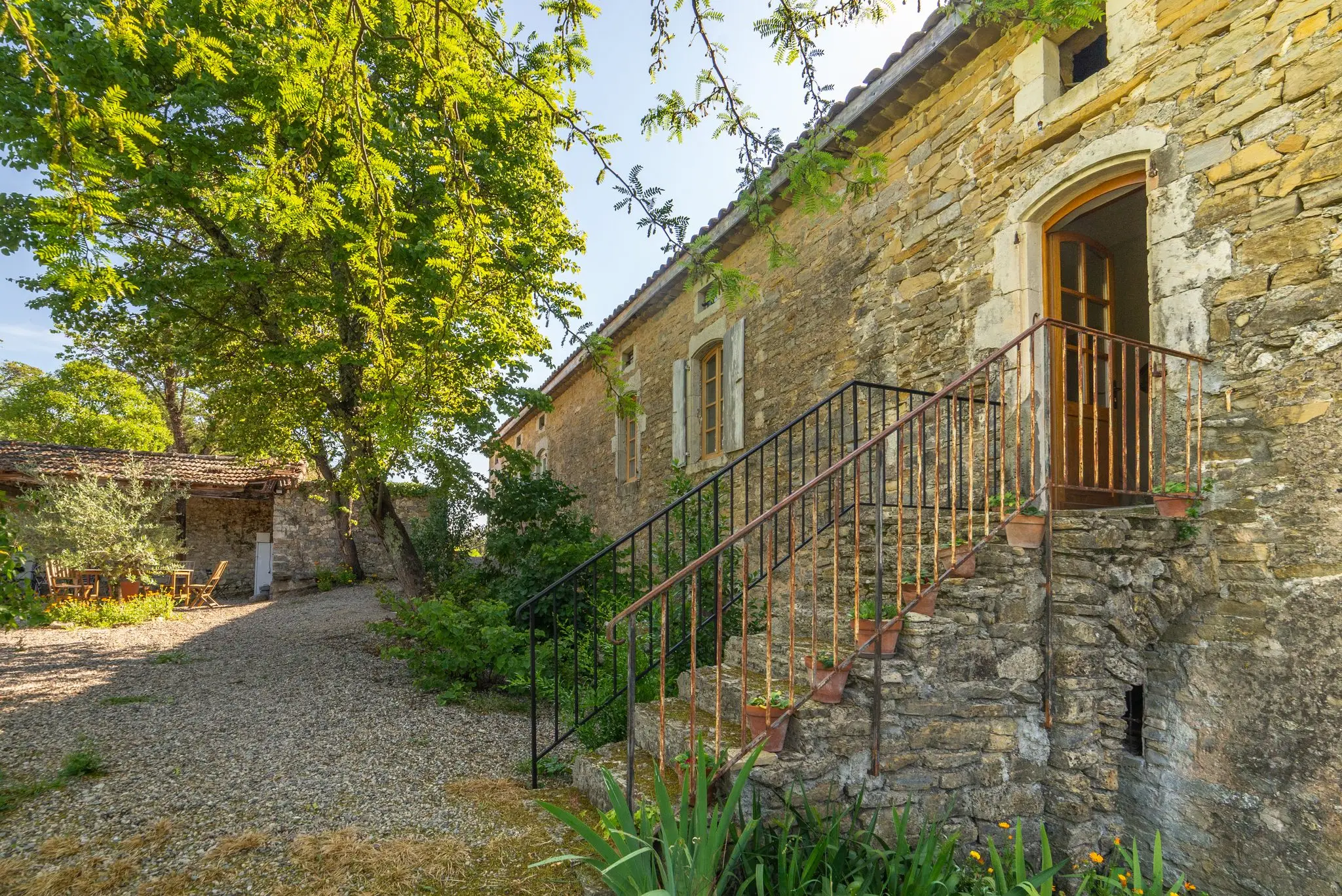 Façade en moellons de pierre d'une maison d'hôtes, escalier extérieur à rampe en fer forgé menant à une porte cintrée, pots de fleurs sur les marches et grands arbres