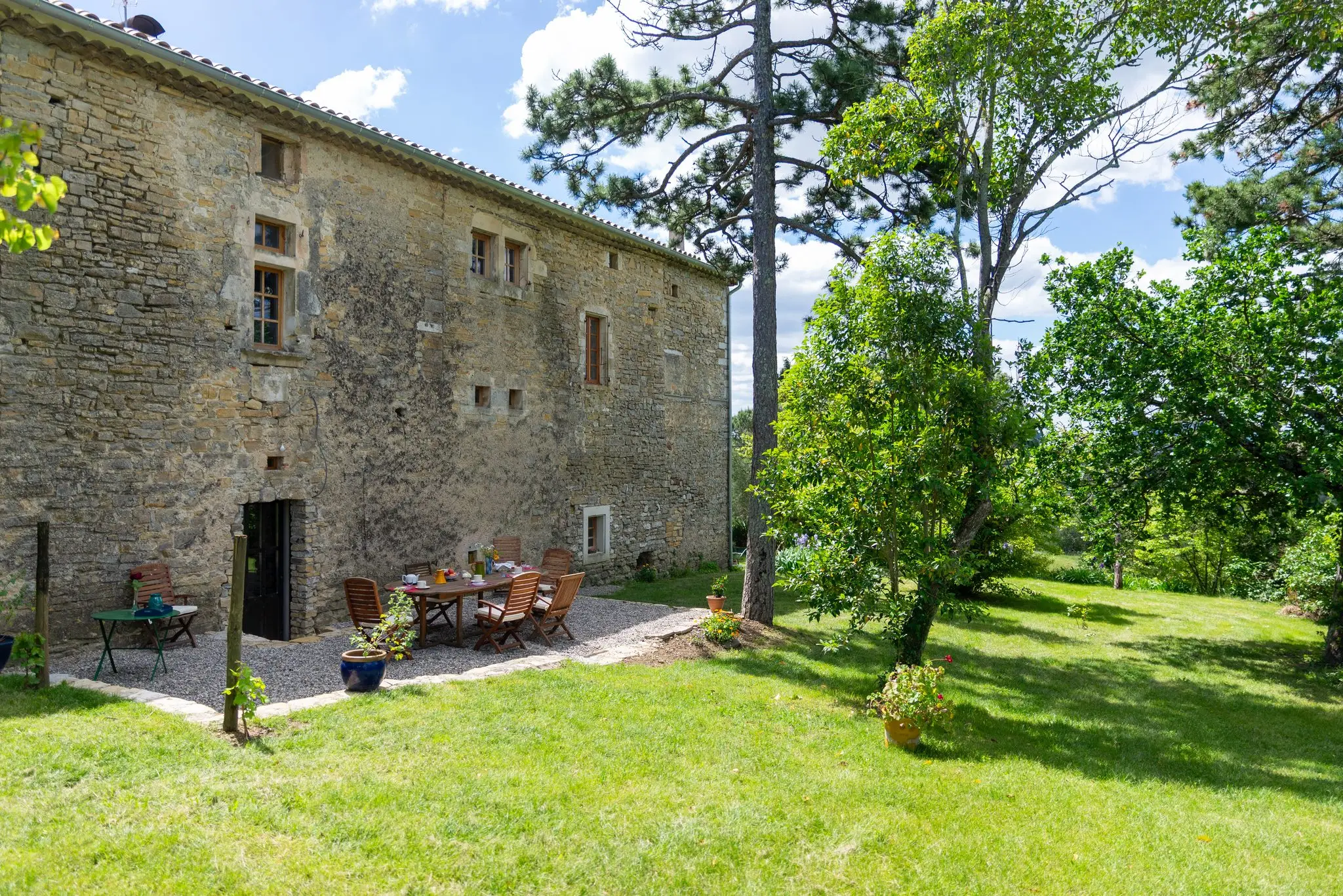 Longue façade en pierre d'une maison d'hôtes ardéchoise, pelouse, pins et table du petit-déjeuner dressée au pied du mur
