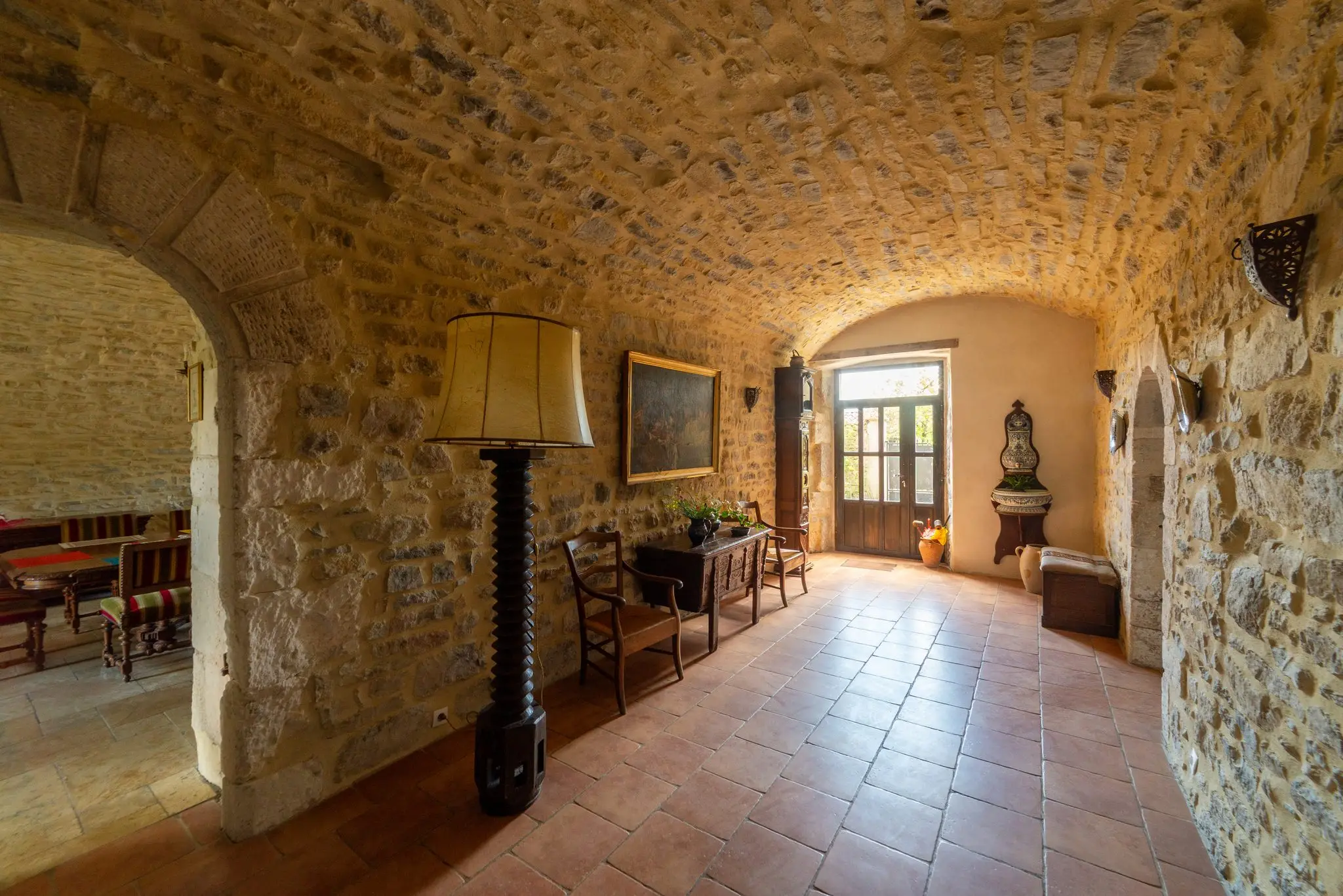 Hall voûté en pierre, sol en tomettes, lampadaire, coffre ancien et tableau, avec une échappée sur la salle à manger par une arche