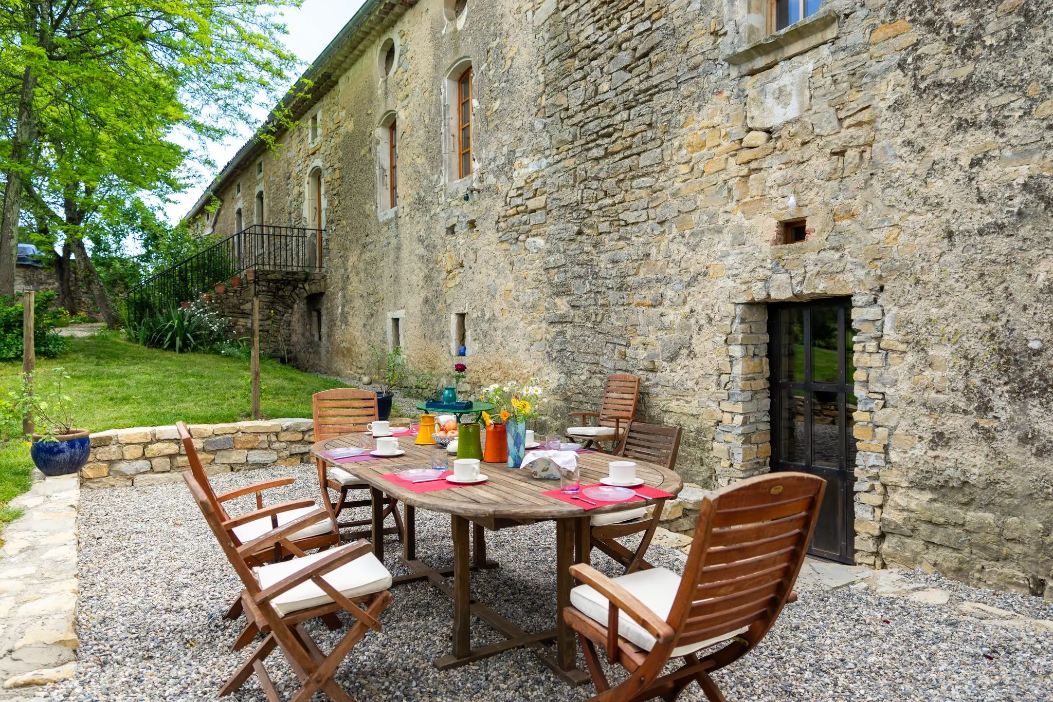 Table en teck dressée pour le petit-déjeuner sur le gravier, devant un mur en pierre et une porte ancienne, pichets colorés et fleurs des champs