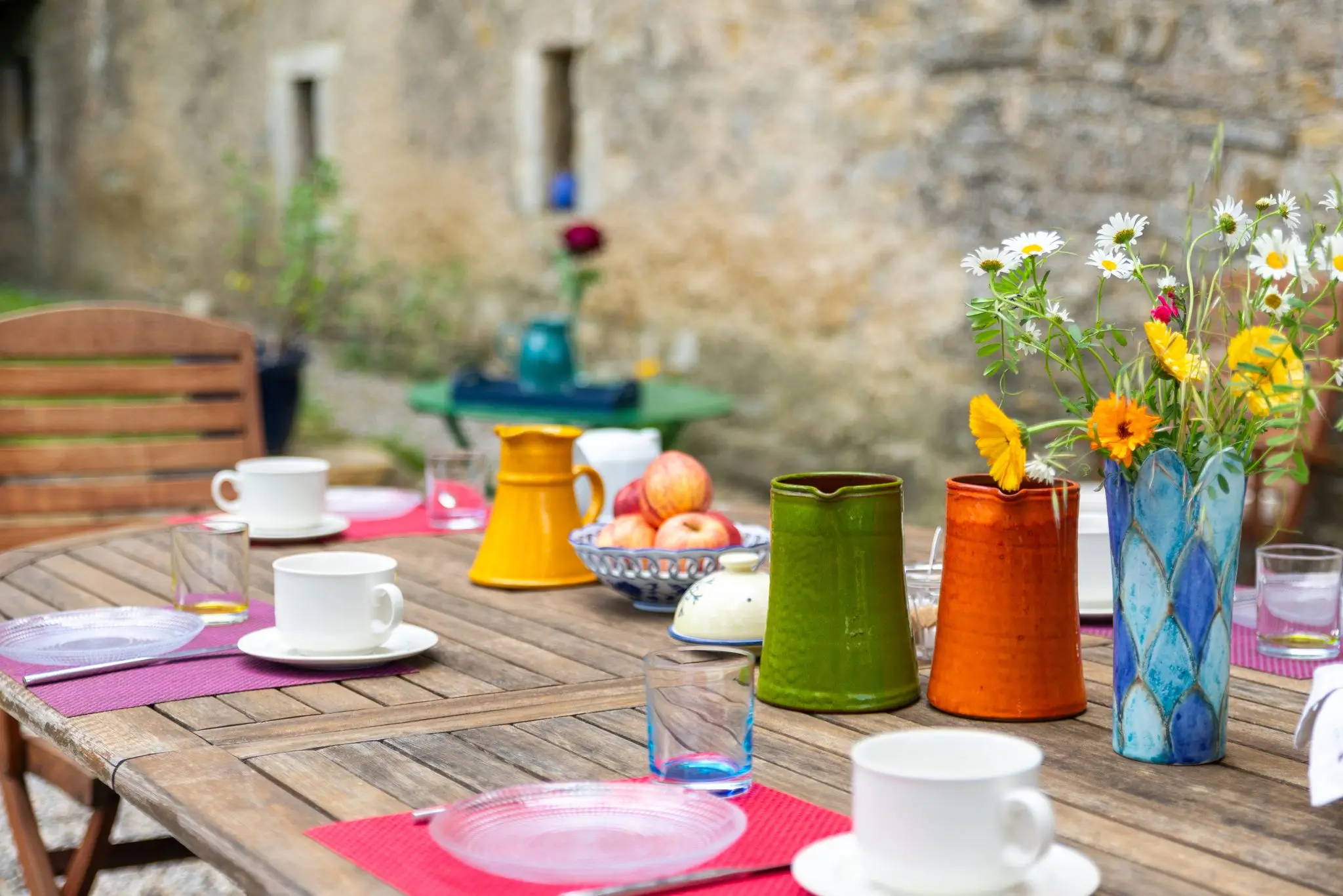 Pichets en céramique jaune, vert et orange, bouquet de marguerites et tasses blanches sur une table en teck, mur en pierre flou à l'arrière