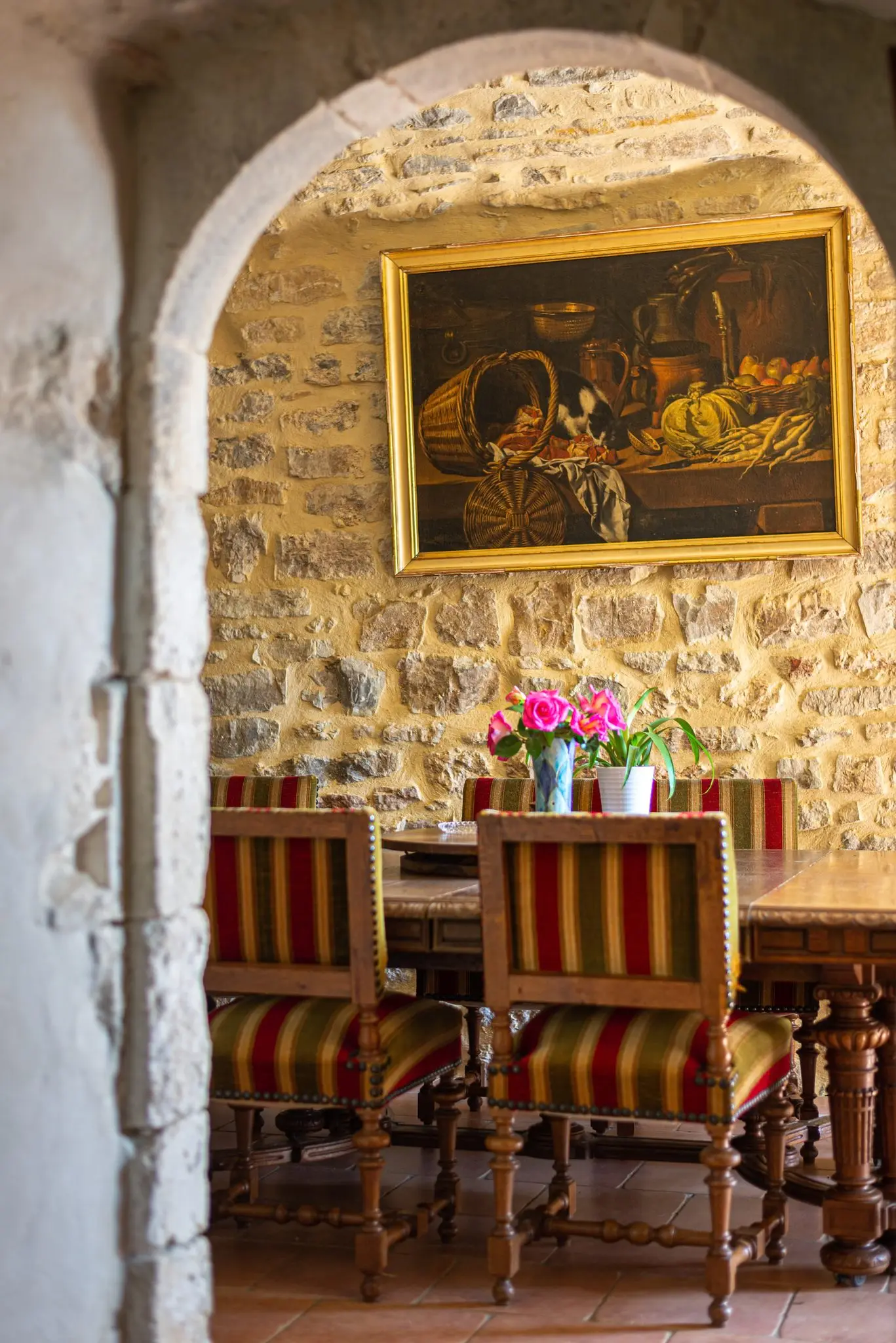 Arcade en pierre ouvrant sur une salle à manger, chaises rayées autour d'une table en bois tourné, bouquet de roses et nature morte encadrée sur le mur de pierre