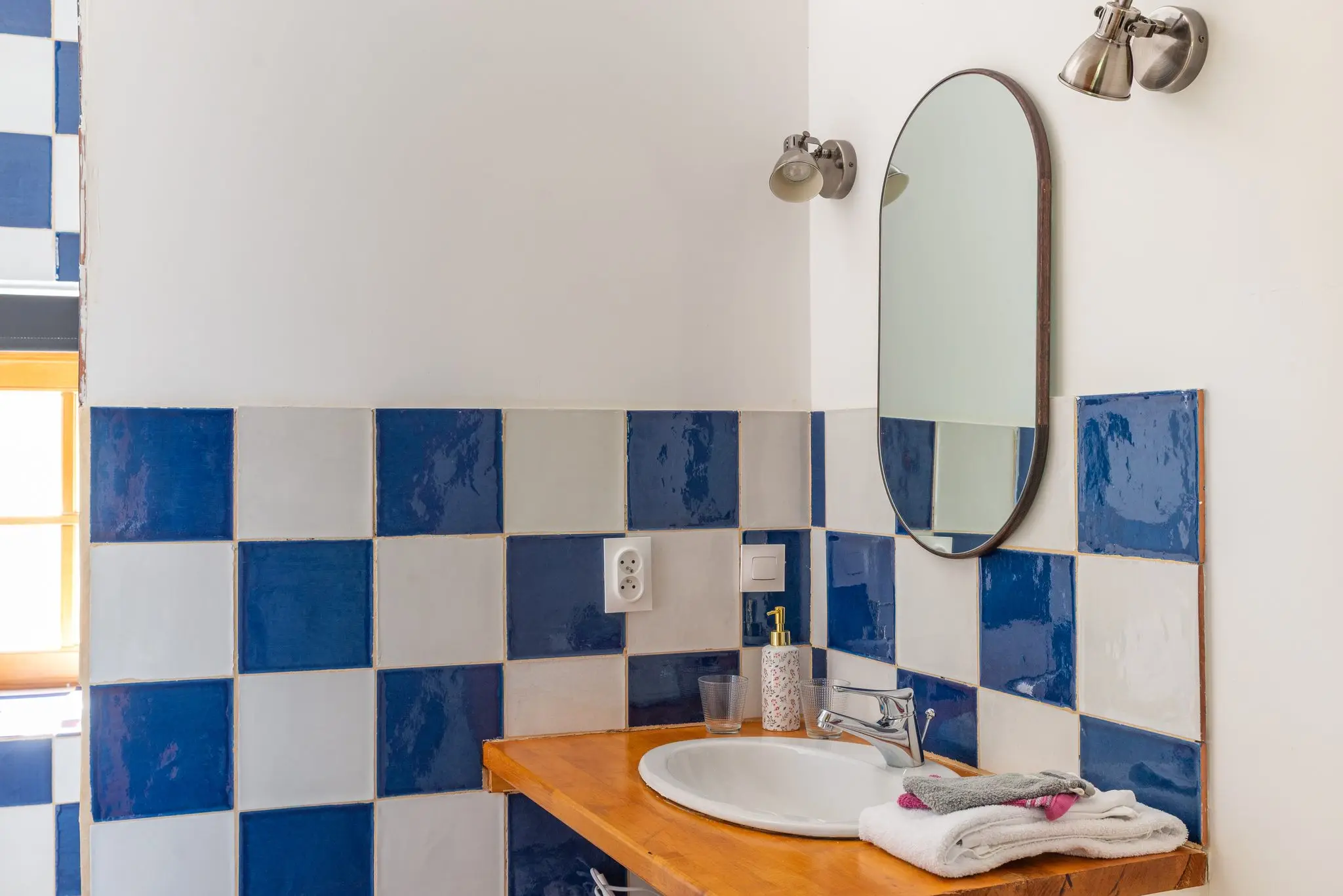 Lavabo sur plan en bois, damier de carreaux émaillés bleus et blancs, miroir ovale et appliques chromées