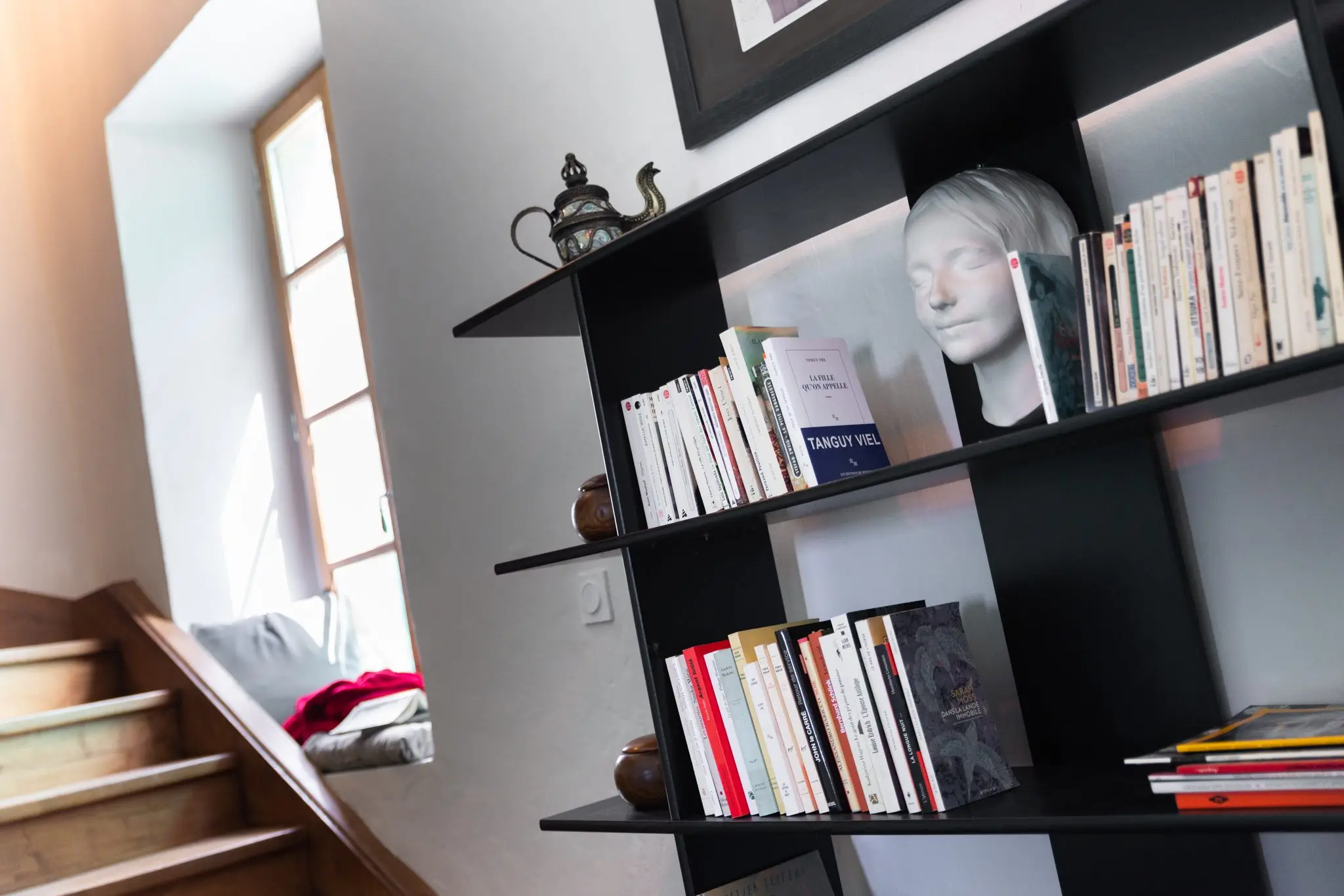 Bibliothèque en métal noir garnie de livres au pied d'un escalier en bois, lumière d'une fenêtre
