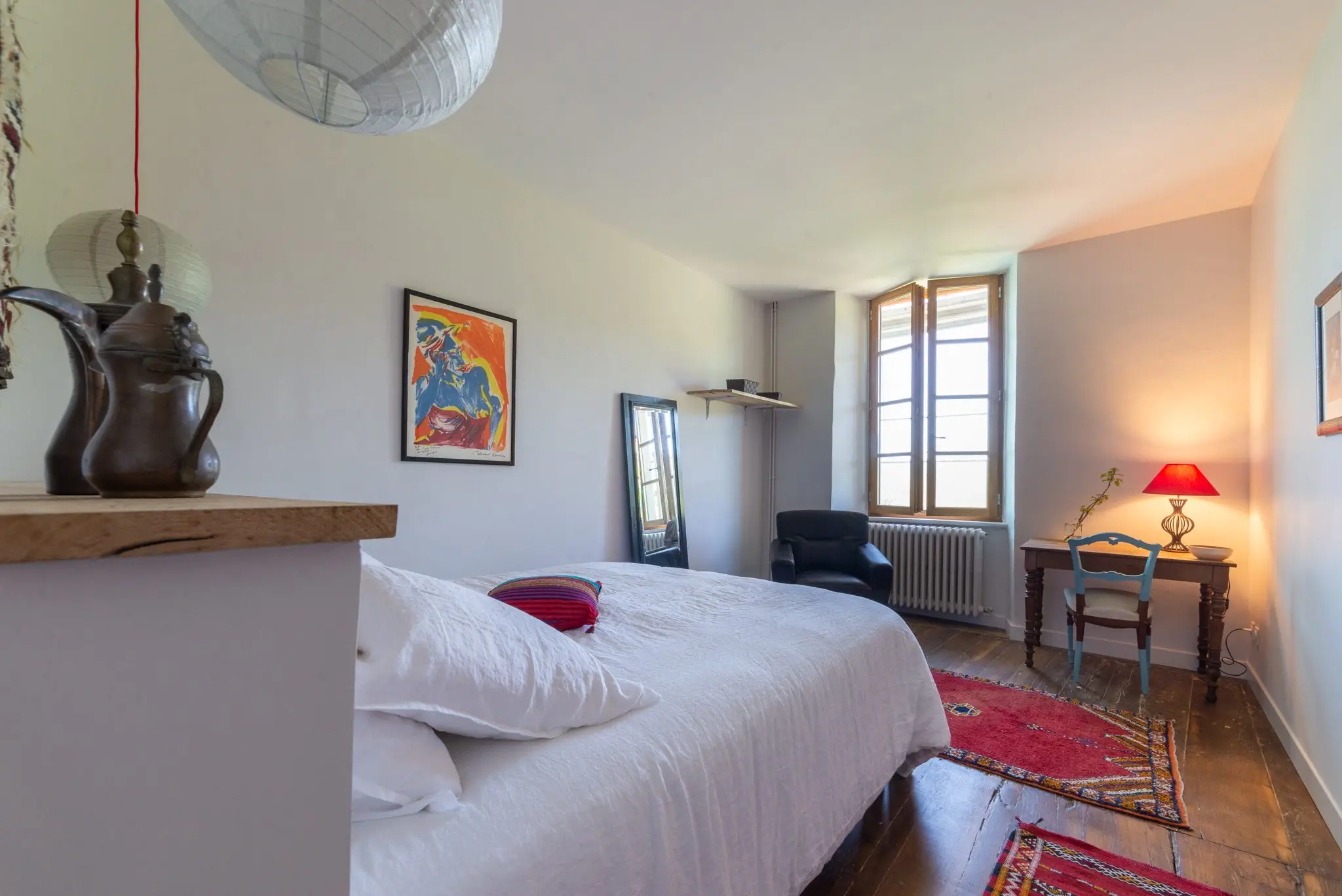 Chambre au lit double blanc, fauteuil en cuir et bureau éclairé par une lampe rouge près de la fenêtre