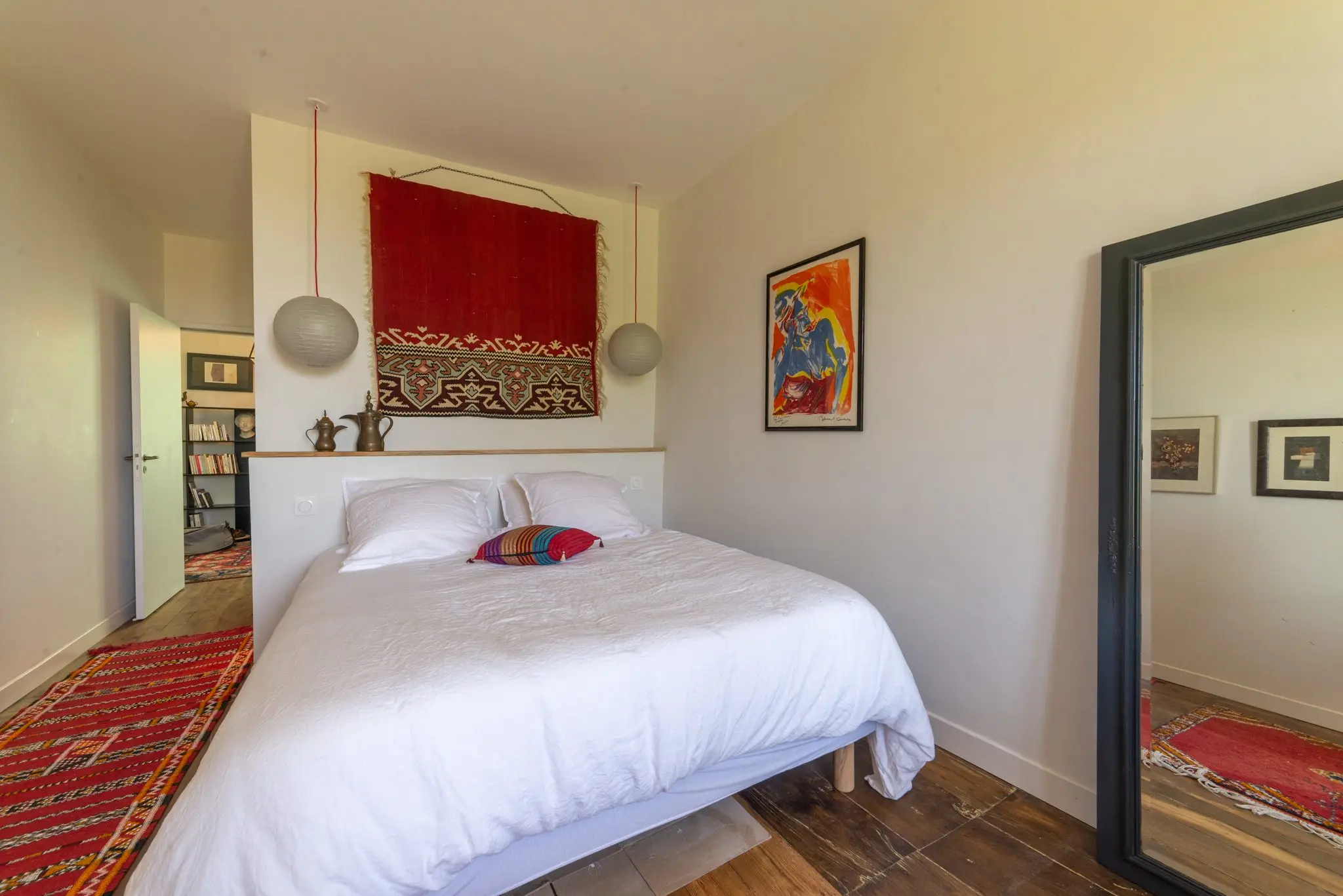 Chambre au lit double blanc sous une tenture rouge brodée, grand miroir et tapis rouge