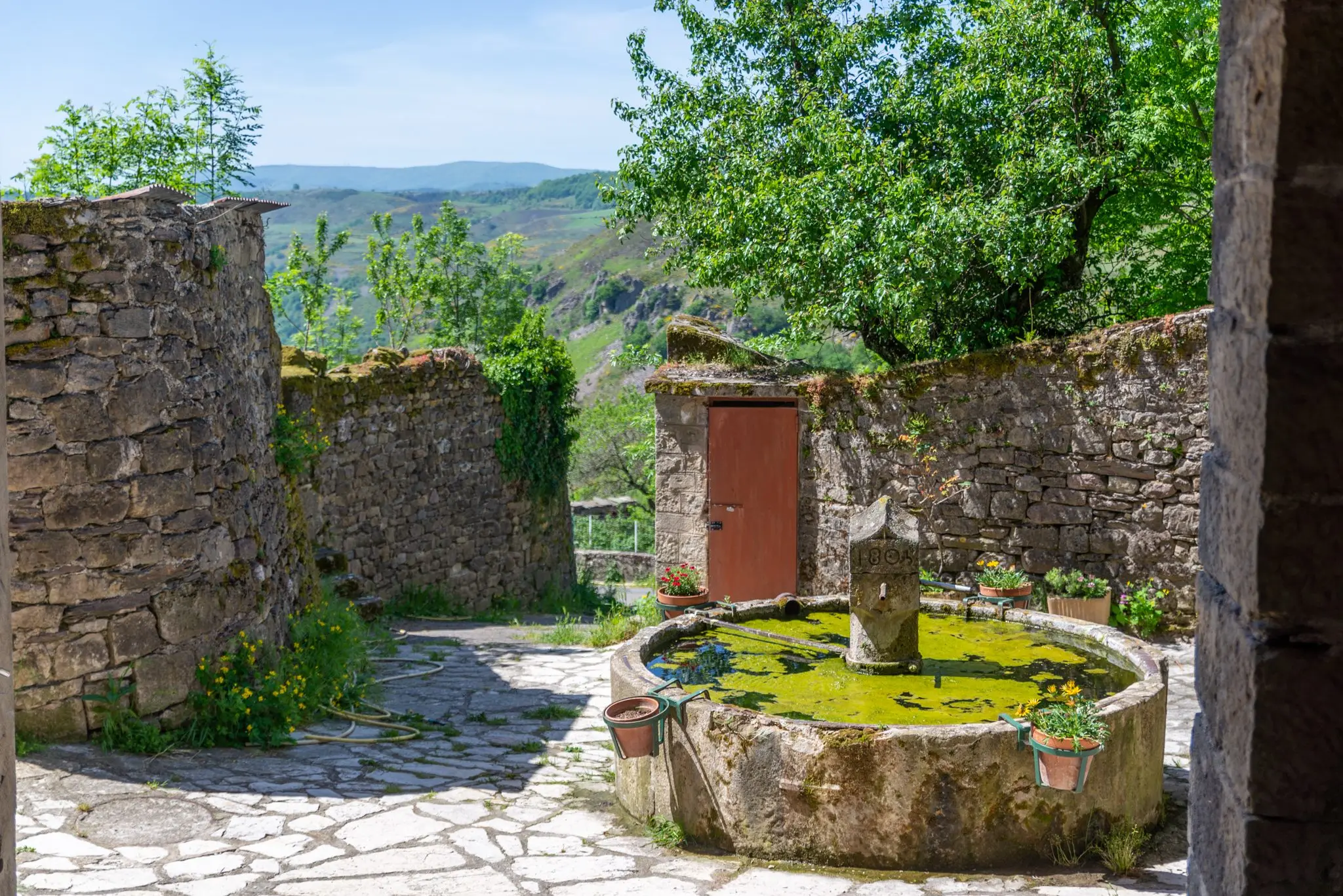 Fontaine ronde en pierre sur une placette dallée entourée de murs en pierre, au cœur d'un village de Lozère