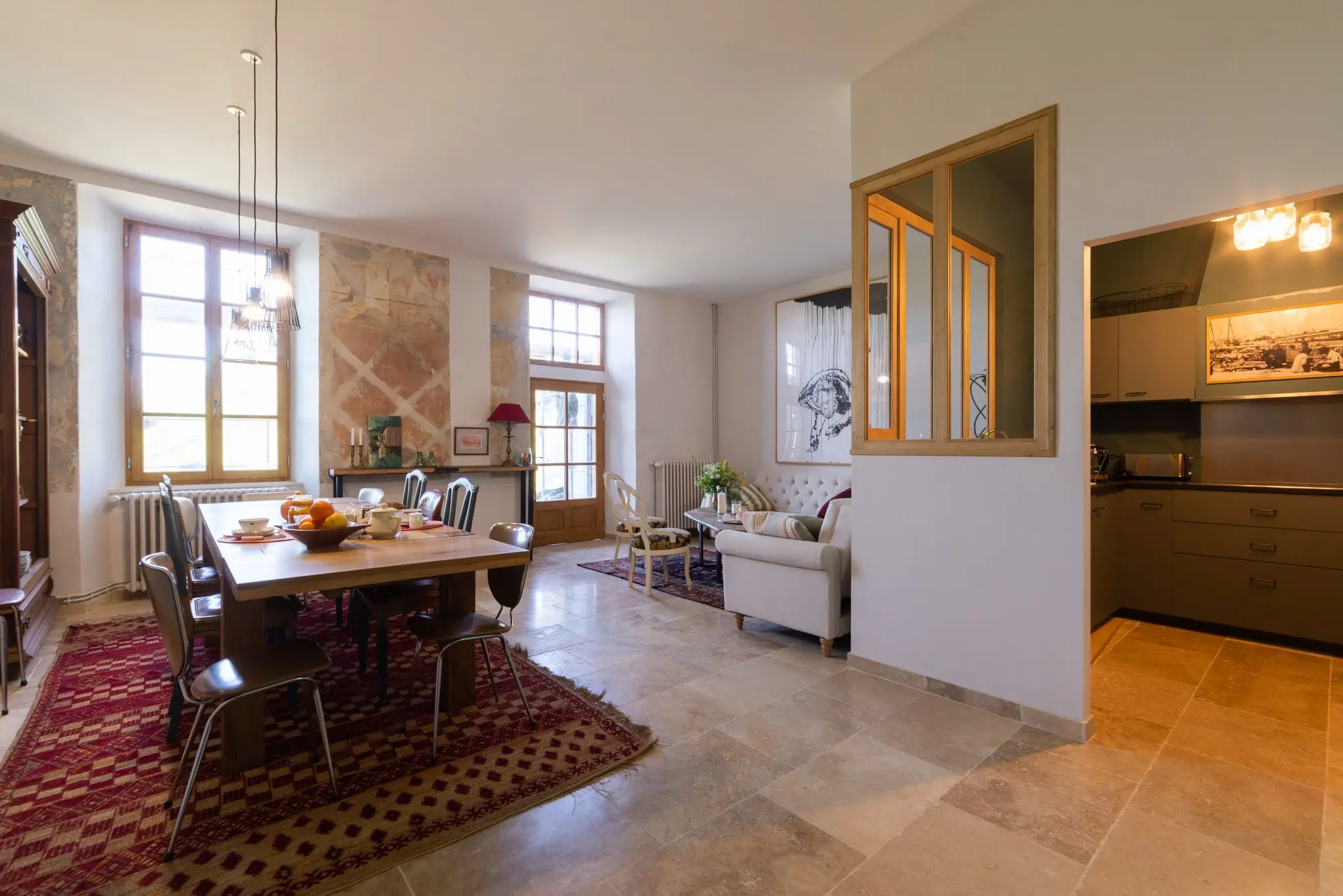 Grande salle aux murs de pierre enduite, table à manger, coin salon et cuisine derrière une verrière