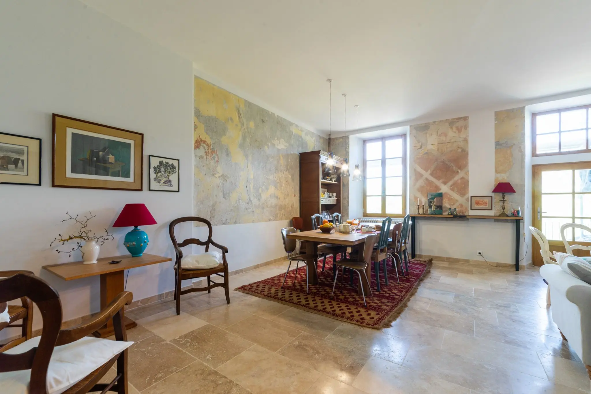 Salle à manger avec longue table, chaises anciennes et tapis rouge, fresque murale et hautes fenêtres