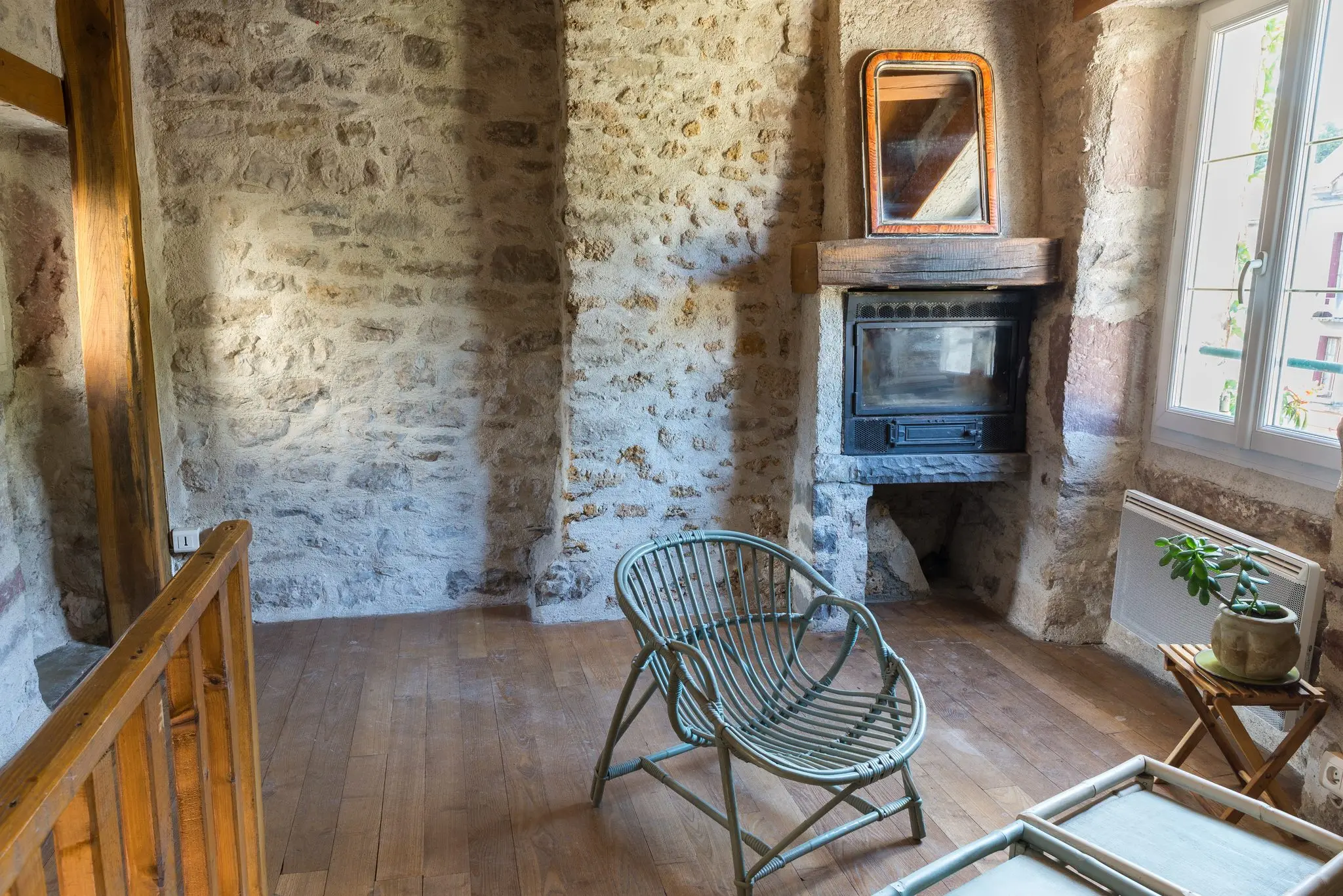 Insert de cheminée et miroir ancien dans un mur en pierre, fauteuil en rotin sur parquet et garde-corps en bois