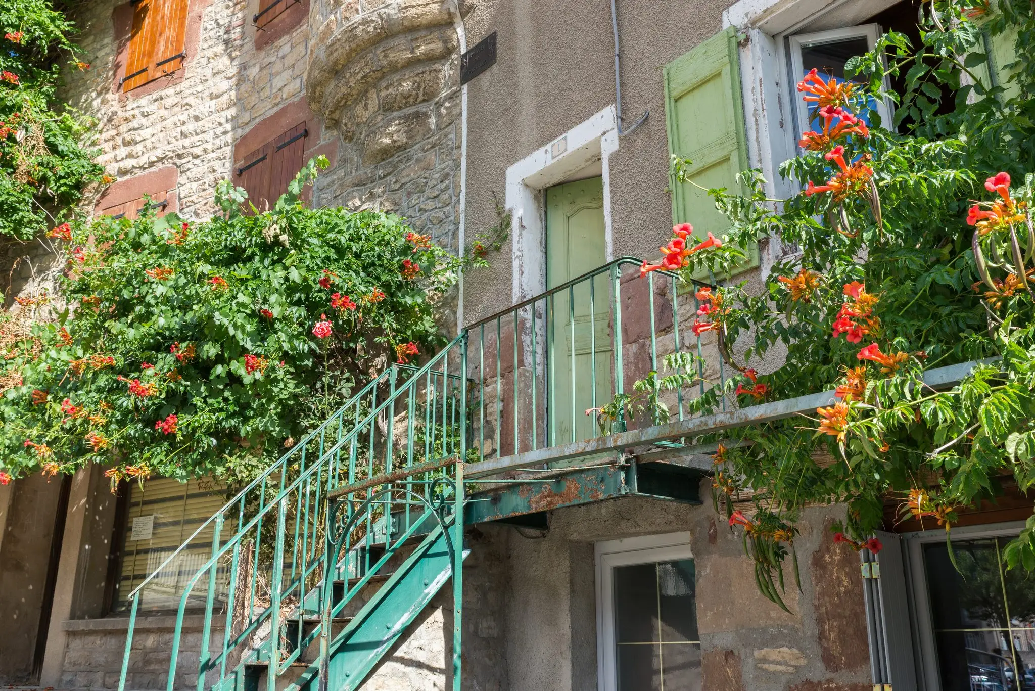 Escalier métallique vert d'eau montant vers une porte aux volets verts, encadré de bignones orange et de murs en pierre