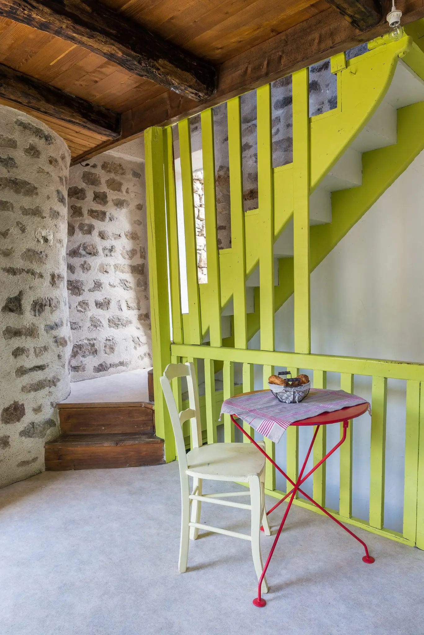 Escalier à garde-corps peint en vert anis, petite table rouge et chaise, mur arrondi en pierre et plafond à solives