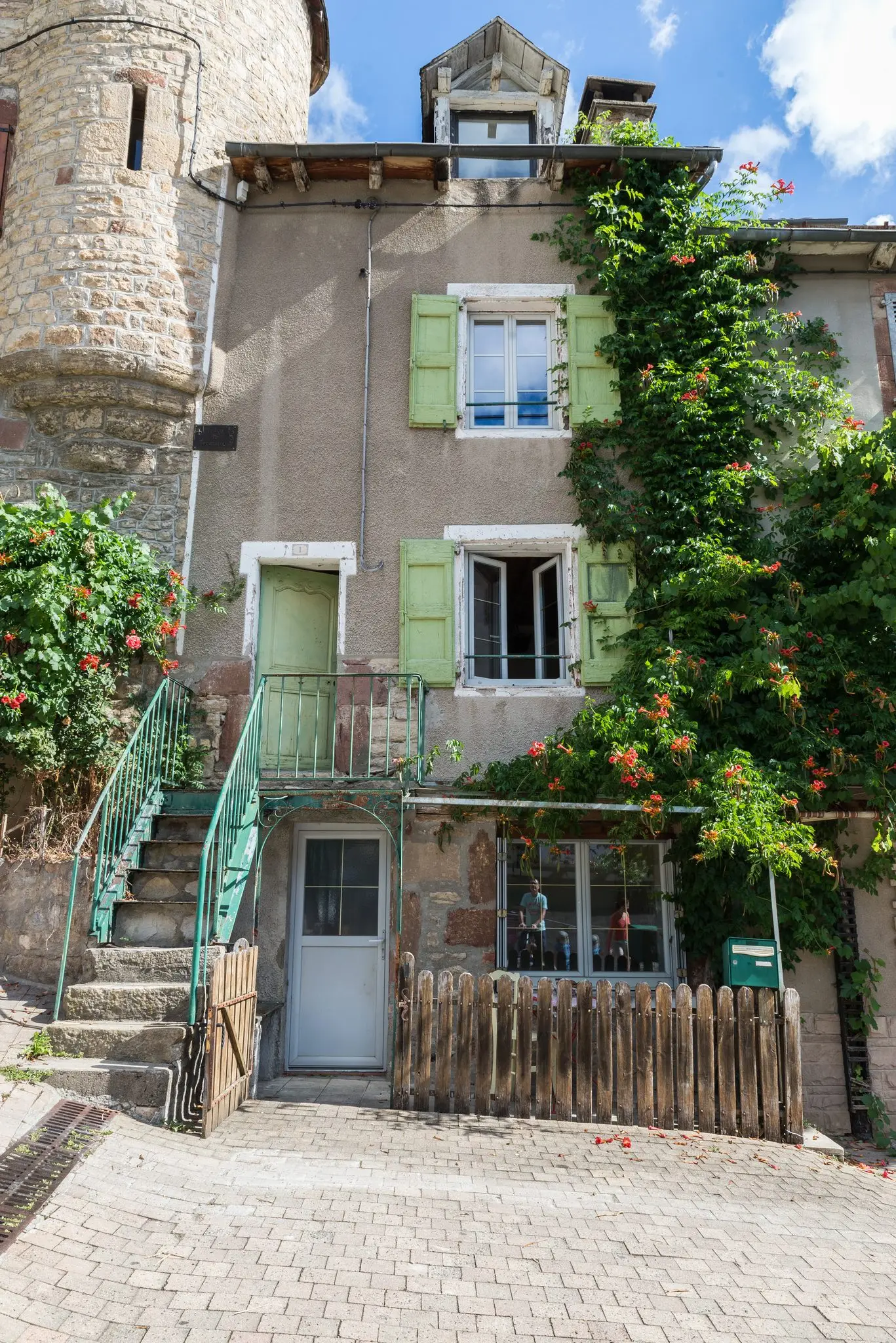 Étroite maison de bourg en pierre enduite, volets verts, escalier extérieur métallique et bignone en fleurs, au pied d'une tour ronde en pierre