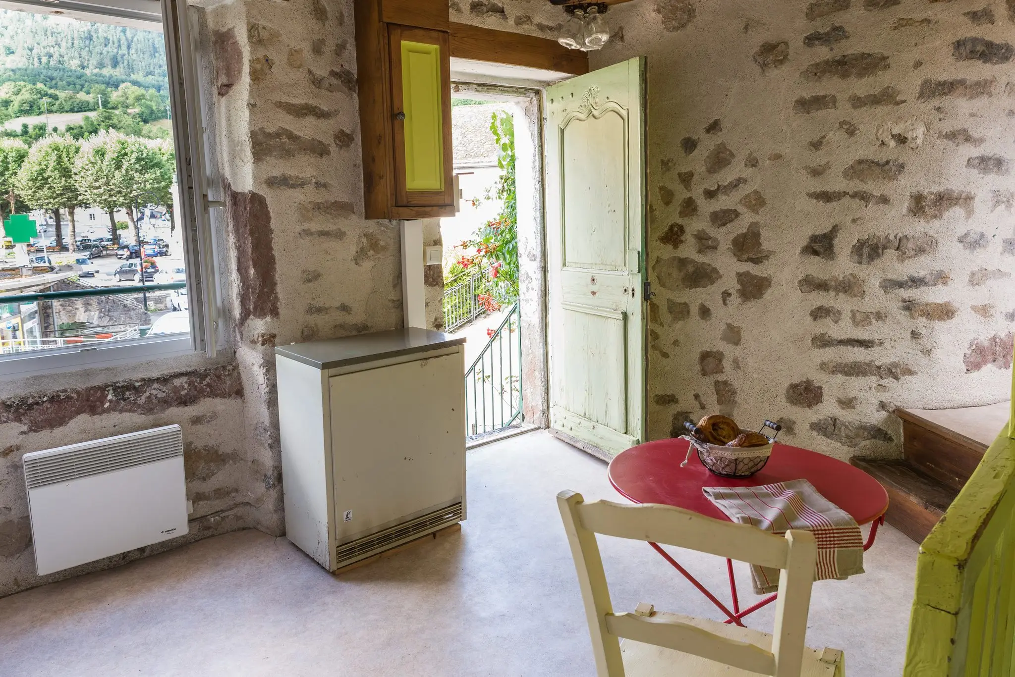 Pièce aux murs en pierre apparente, coin cuisine, table ronde rouge et porte ancienne ouverte sur un balcon fleuri