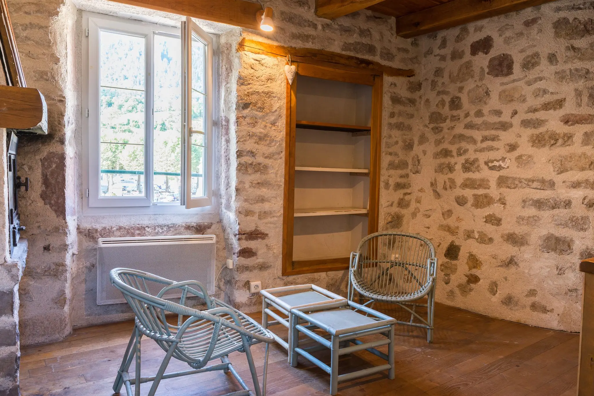 Séjour au parquet ancien et murs en pierre apparente, niche-bibliothèque en bois, fauteuils en rotin et fenêtre donnant sur la montagne