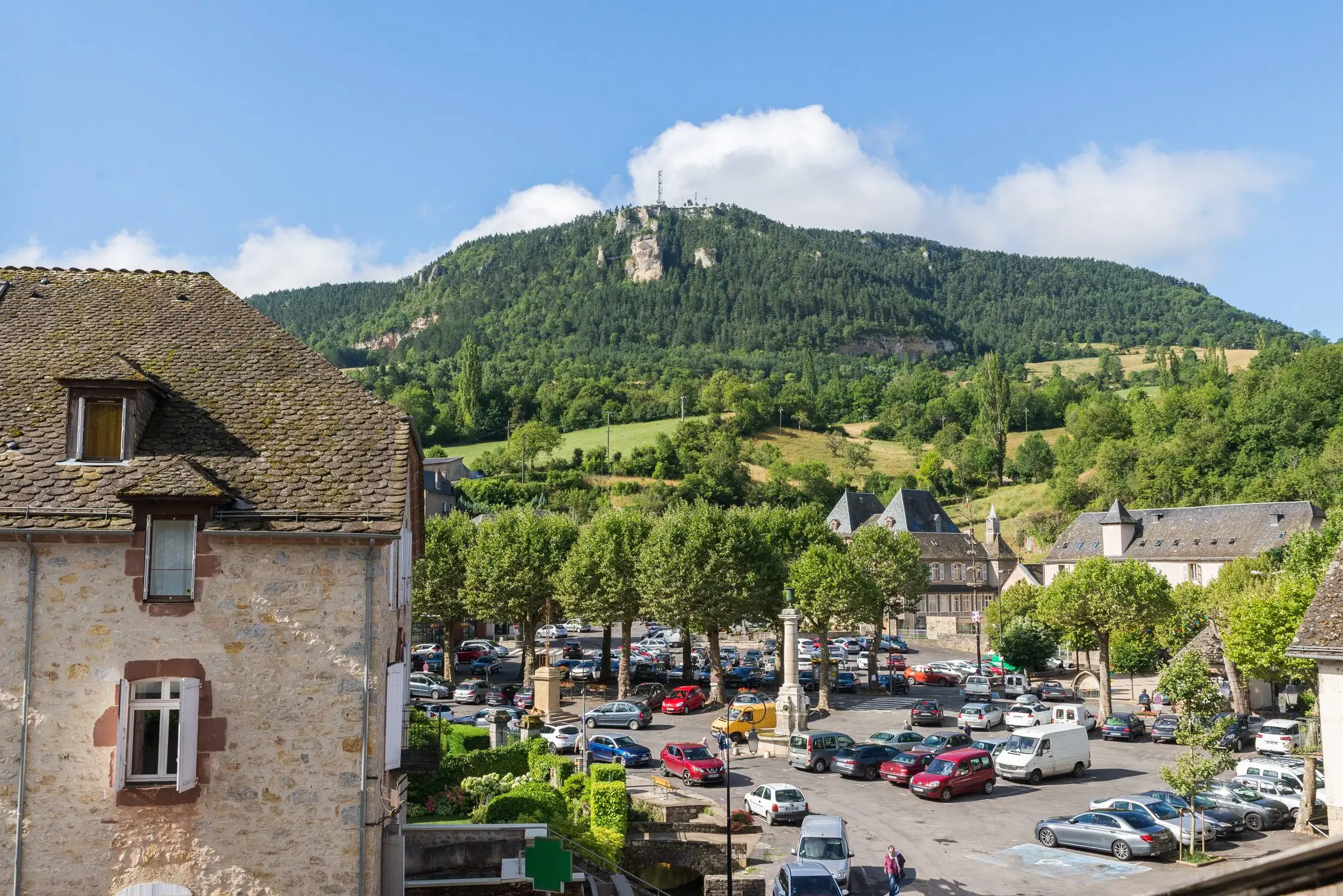 Place de bourg plantée d'arbres au pied d'une montagne boisée surmontée de falaises calcaires, toits en lauze au premier plan