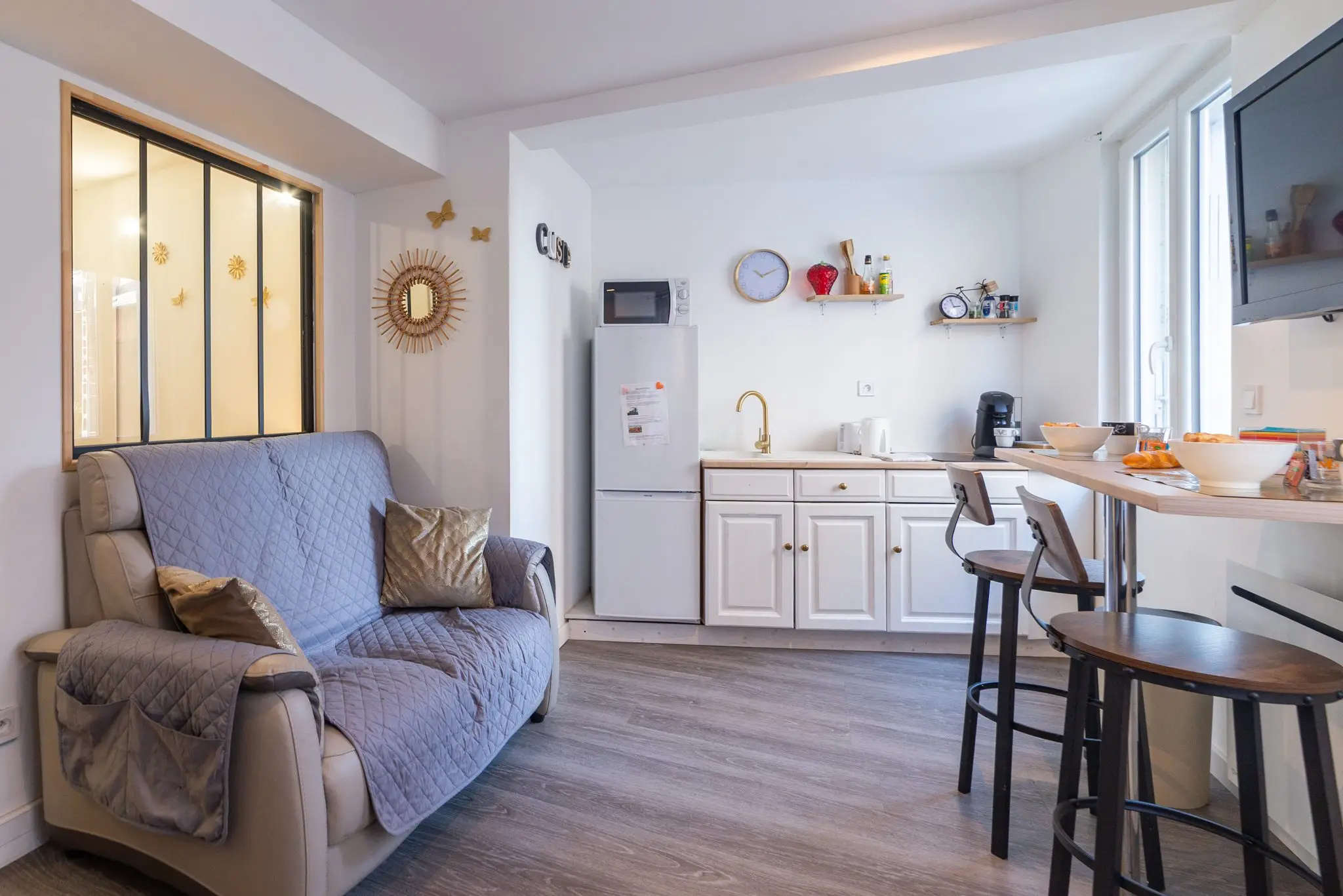 Séjour d'un studio meublé, canapé gris, coin cuisine blanc et bar avec tabourets, verrière au fond